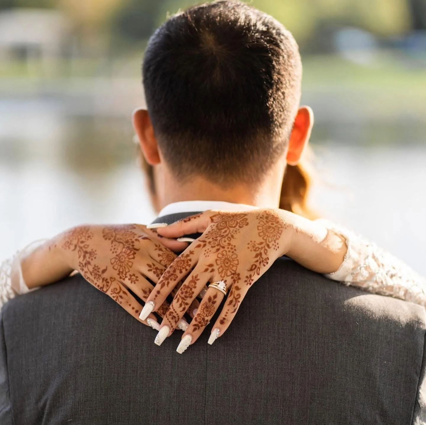 A bride with henna-adorned hands embracing a groom in a suit, viewed from behind near a water body with trees in the background.