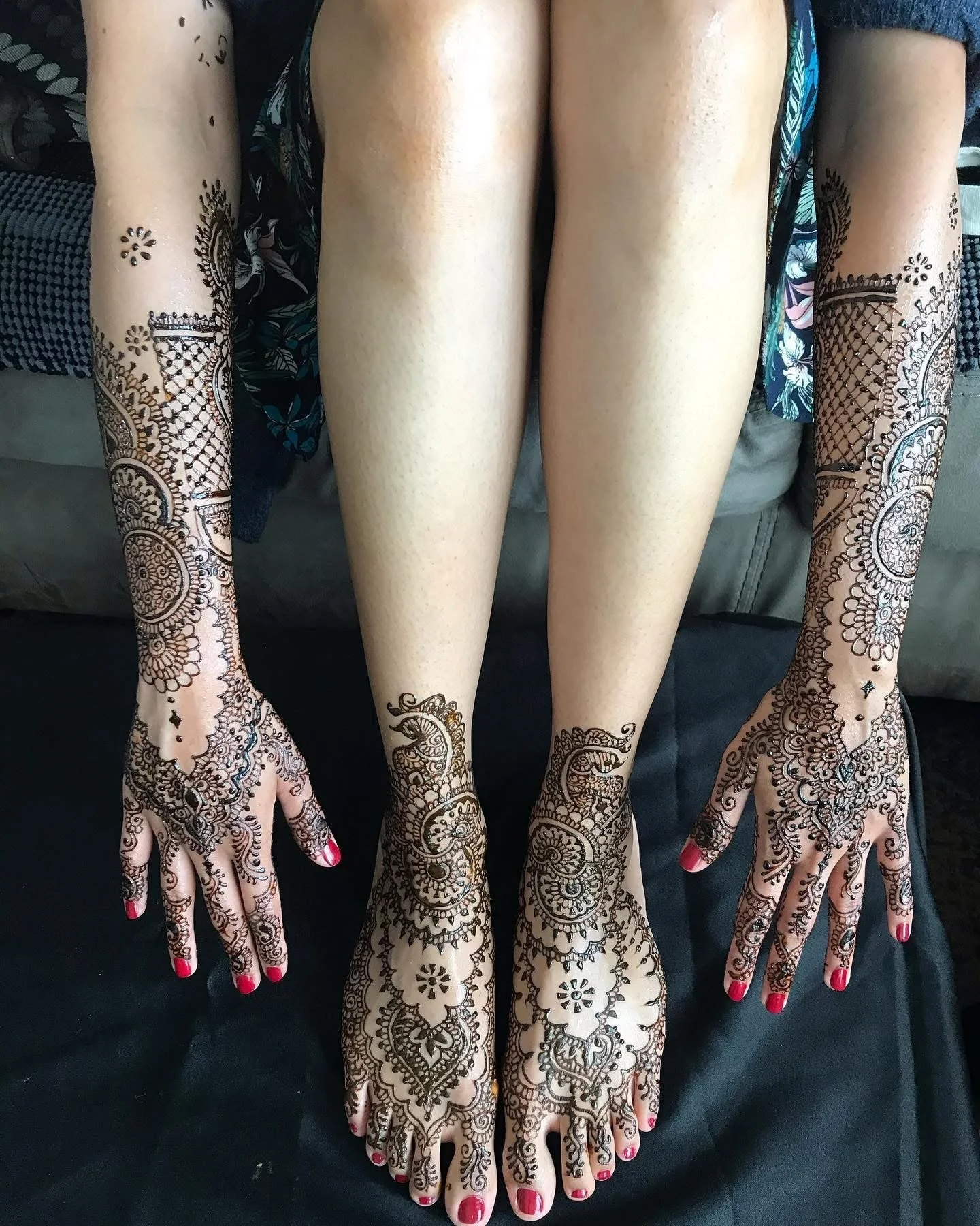 Person with detailed henna designs on hands, arms, and feet, sitting with legs extended and hands resting on the ground.
