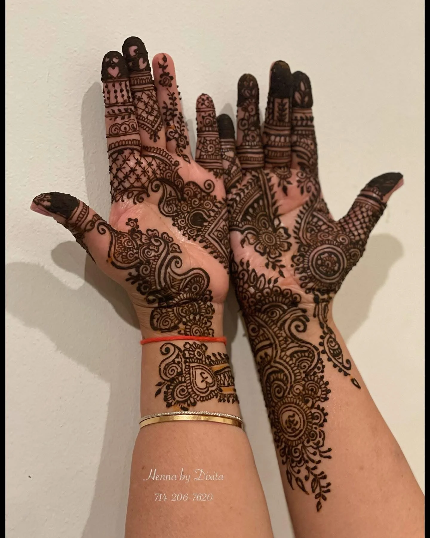 Hands adorned with intricate henna designs, featuring floral and geometric patterns, against a plain wall background.
