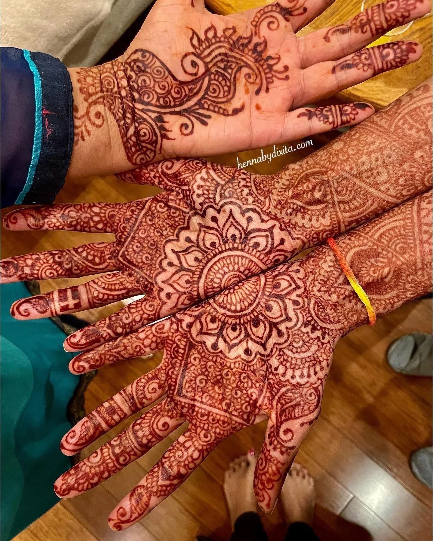 Two hands decorated with intricate henna designs, featuring floral, paisley, and geometric patterns, with a wooden floor and feet visible in the background.