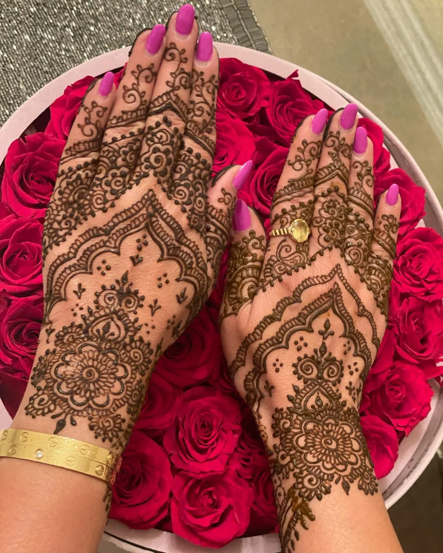 Hands with intricate henna designs, pink nail polish, gold bracelet, and a ring, resting on a bed of pink roses.