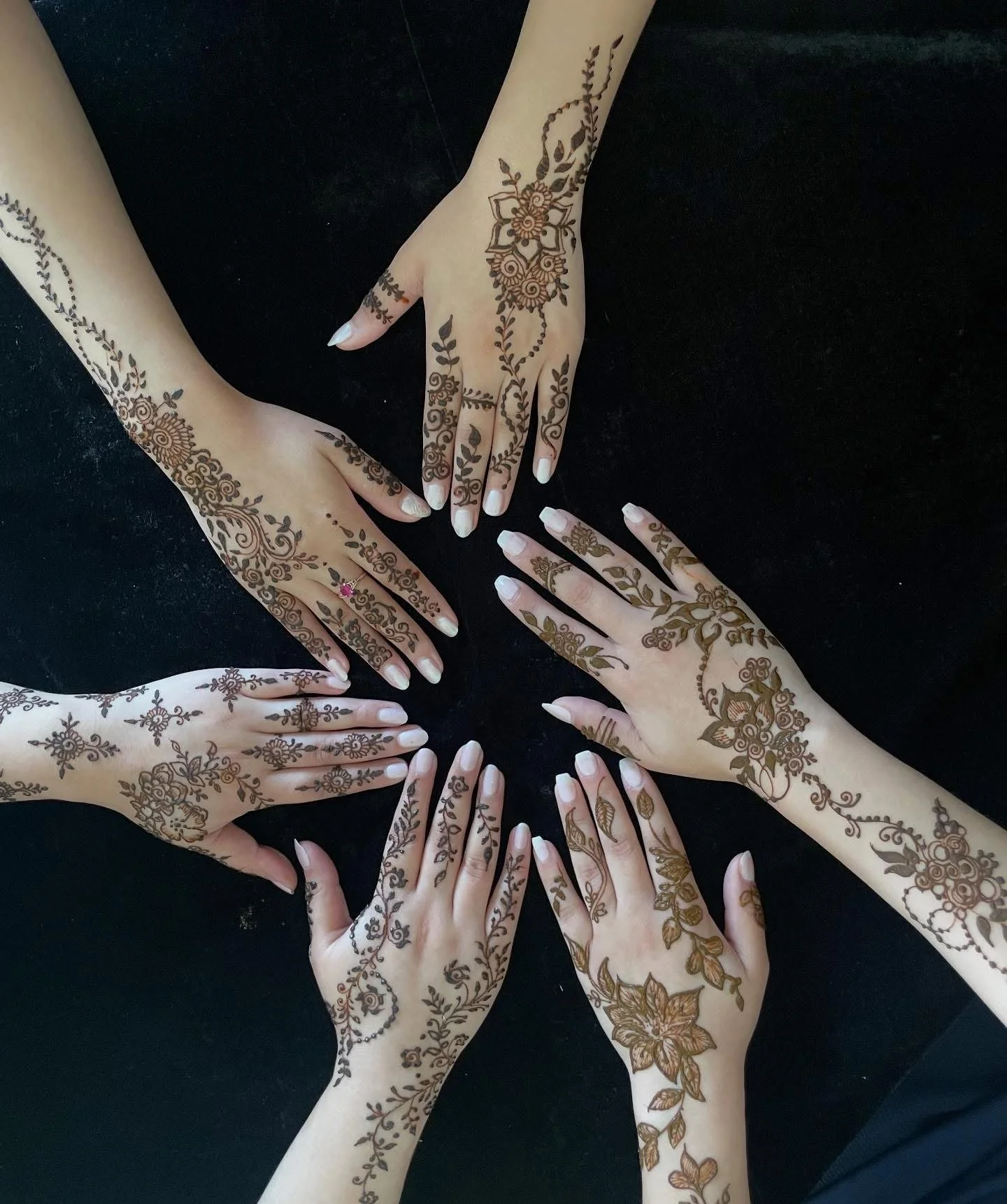 Four hands with intricate henna designs placed together on a black surface.
