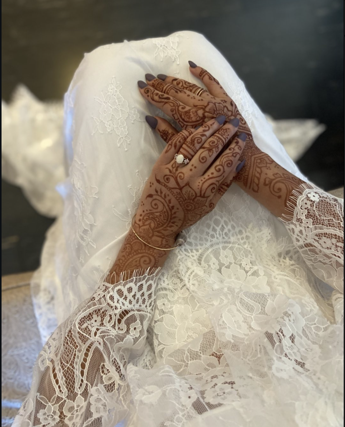 Close-up of a woman with intricately henna-dyed hands and wearing jewelry, dressed in a white lace dress, sitting with her hands clasped on her lap.
