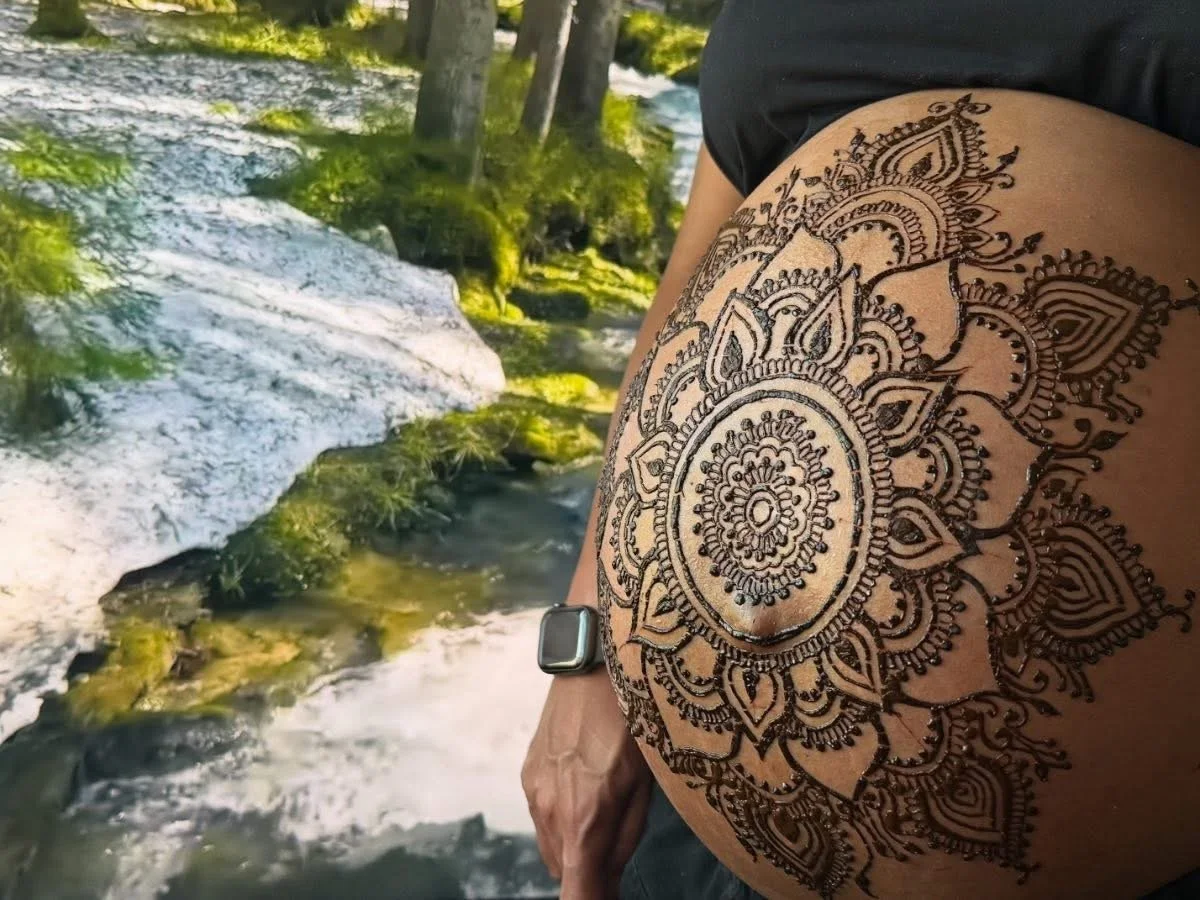 Close-up of a person's thigh with an intricate mandala henna tattoo, outdoors near a flowing stream with trees and moss-covered rocks in the background.