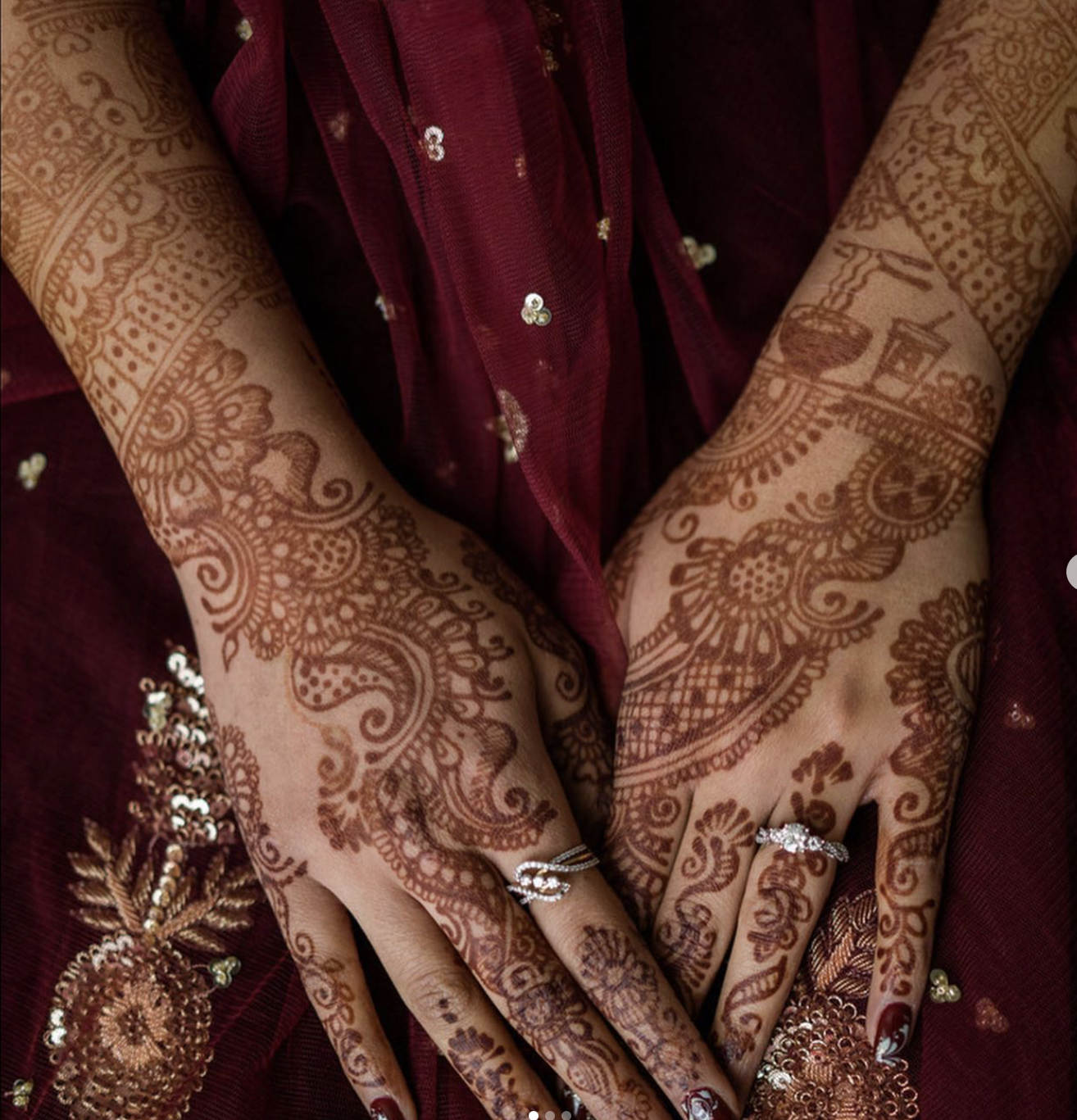 Close-up of hands adorned with intricate henna designs, wearing rings, and set against a maroon and gold traditional dress with embroidery.
