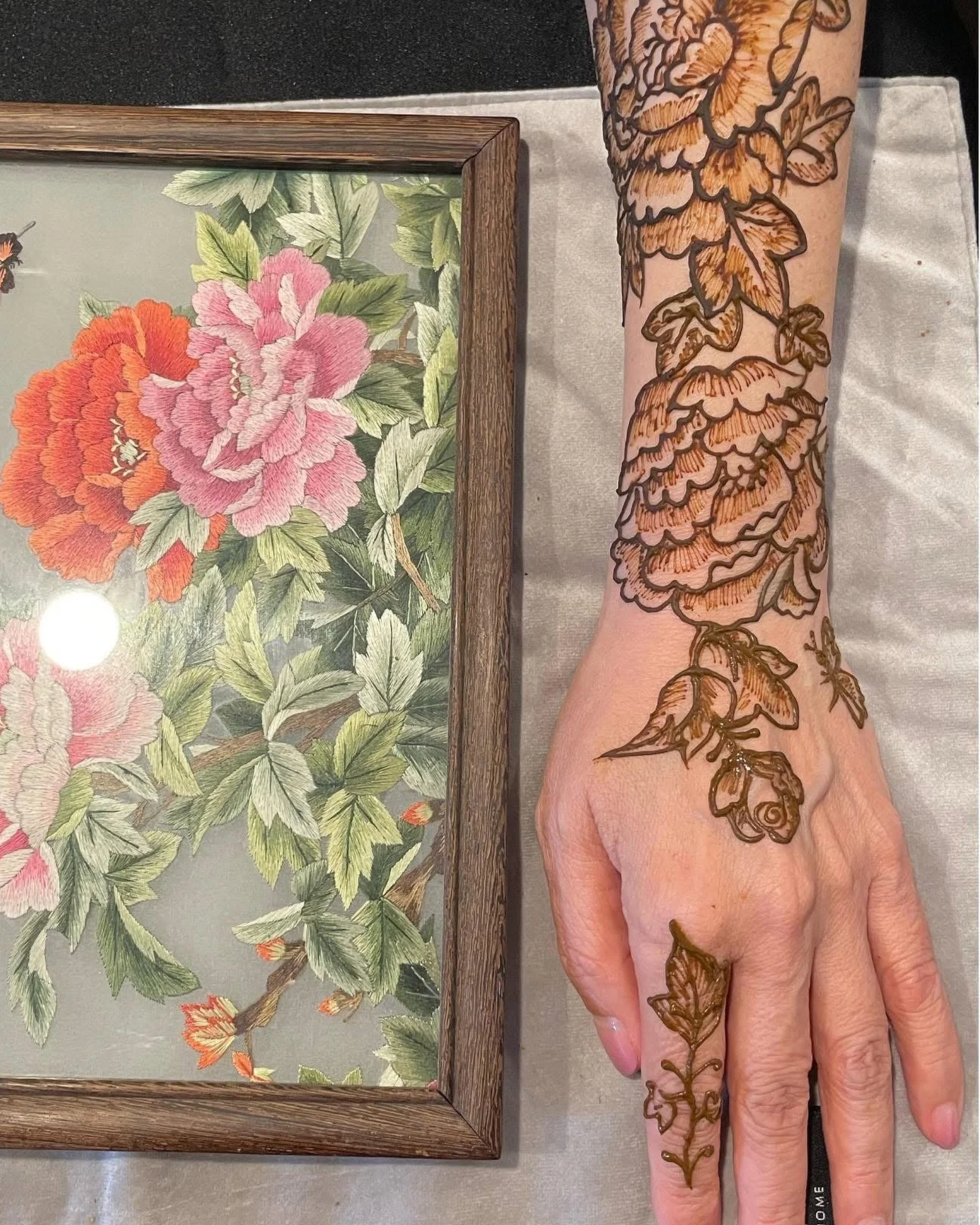 Forearm with intricate henna floral design next to a framed botanical print of pink and orange peonies and green leaves.
