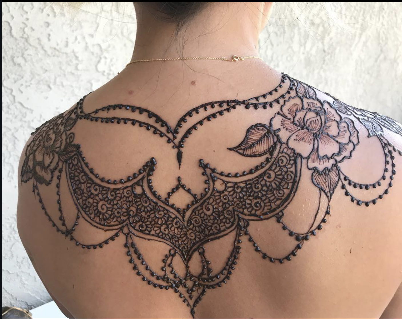 A person's upper back and shoulder with an intricate black tattoo design, featuring floral elements, beads, and lace-like patterns.