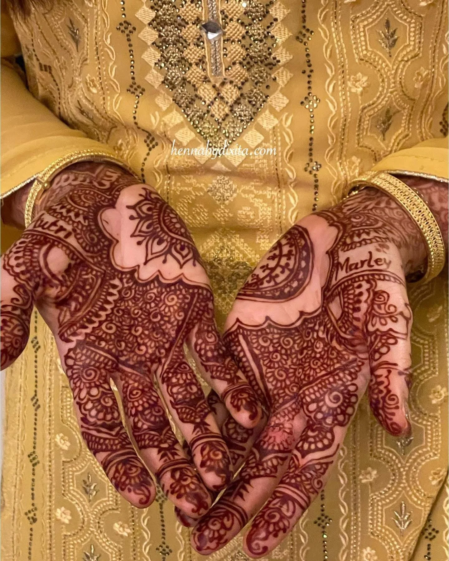 Close-up of hands with intricate henna designs, wearing traditional yellow and gold embroidered attire.