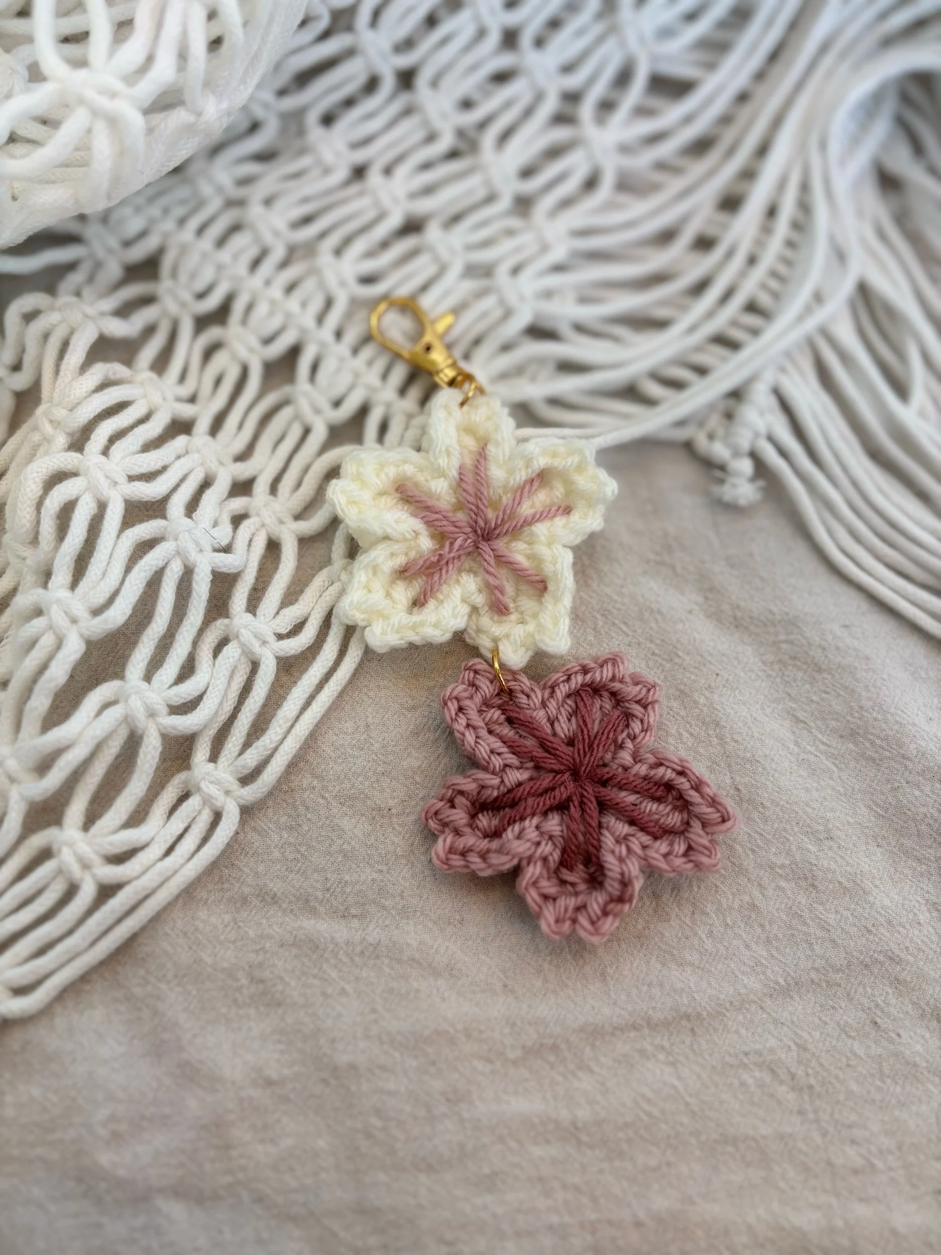 A pair of crocheted flower-shaped charms, one cream-colored with pink accents and one pink with darker pink accents, attached to a gold clasp, lying on a beige fabric surface with a white macrame background.
