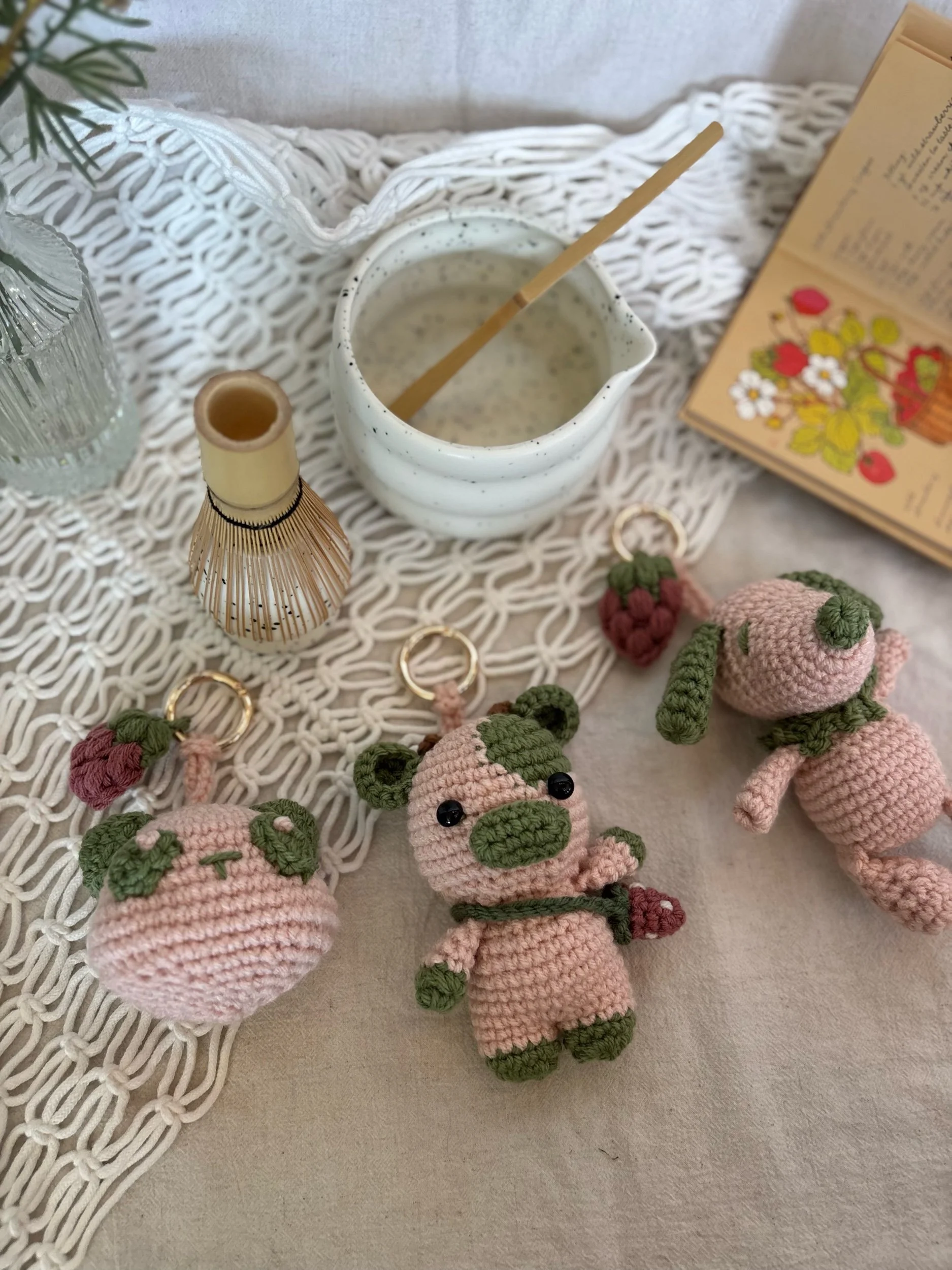 Collection of crocheted animal keychains including a bear, a pig, and a rabbit, arranged on a cream tablecloth alongside a bowl of cream, a glass of water, a reed diffuser, a small open notebook, and a plant in the background.