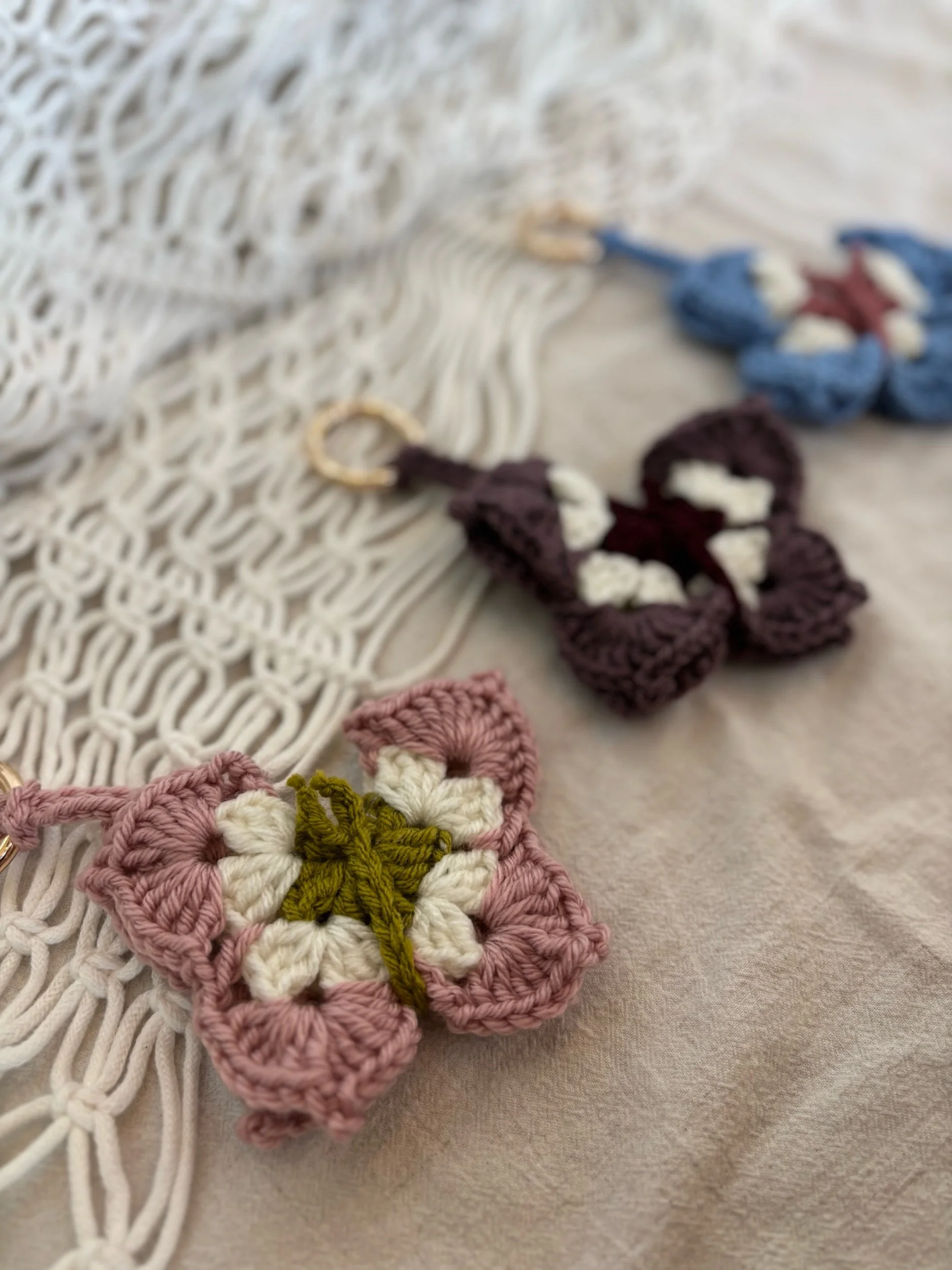 Close-up of handmade crochet flower keychains in pink, purple, and blue colors, laid on a beige fabric with a white macrame hanging on the side.