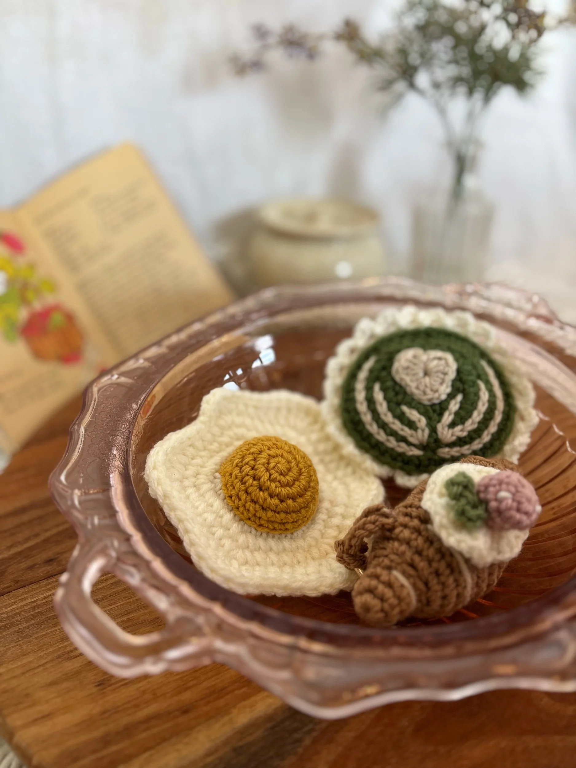 A glass bowl containing three crocheted food items resembling a fried egg, a sushi roll, and a small sushi with a flower topping, placed on a wooden surface with a blurred background of an openbook, a jar, and flowers in a vase.
