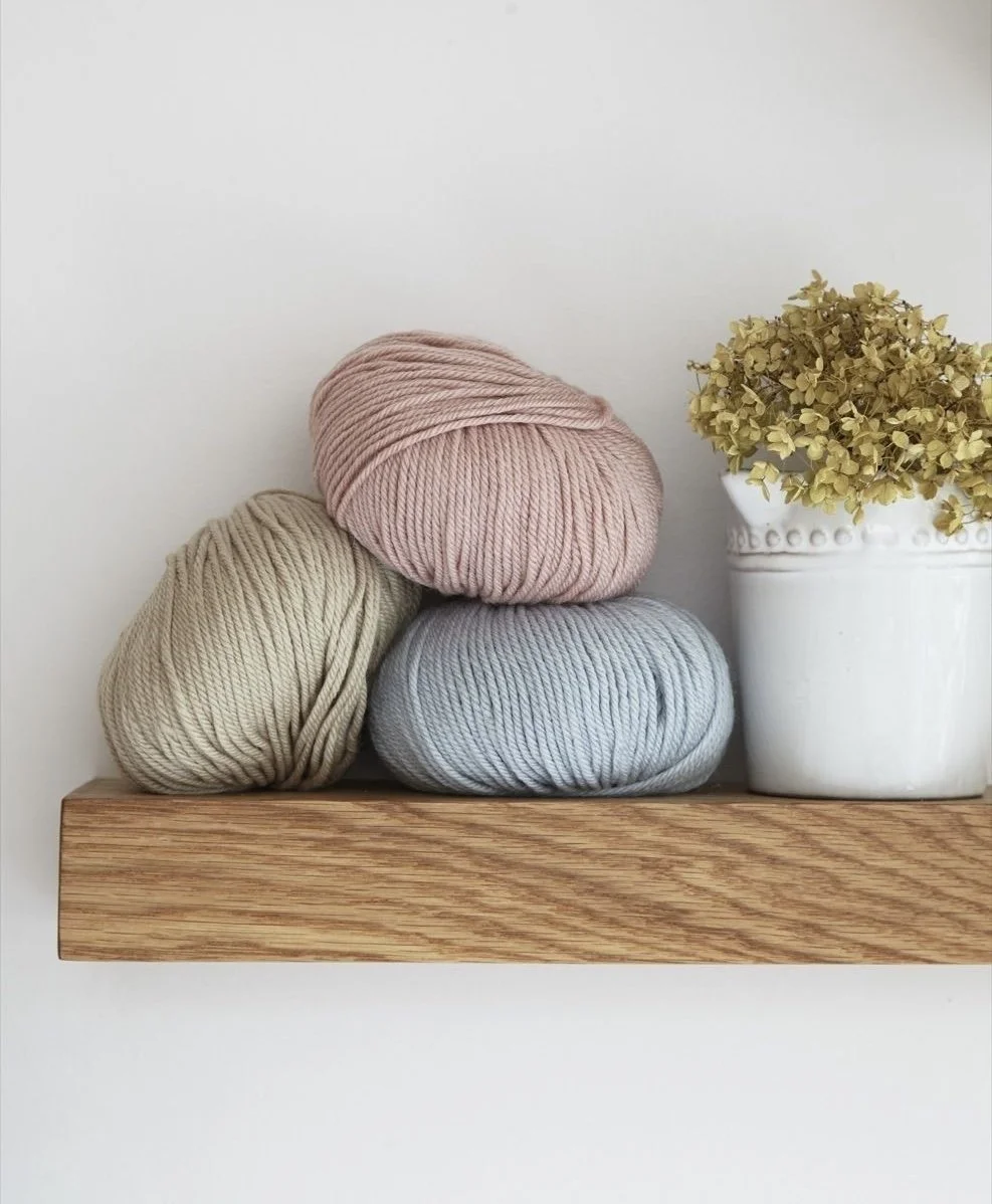 Three skeins of yarn in beige, pink, and gray stacked on a wooden shelf next to a white vase with dried yellow hydrangea flowers.