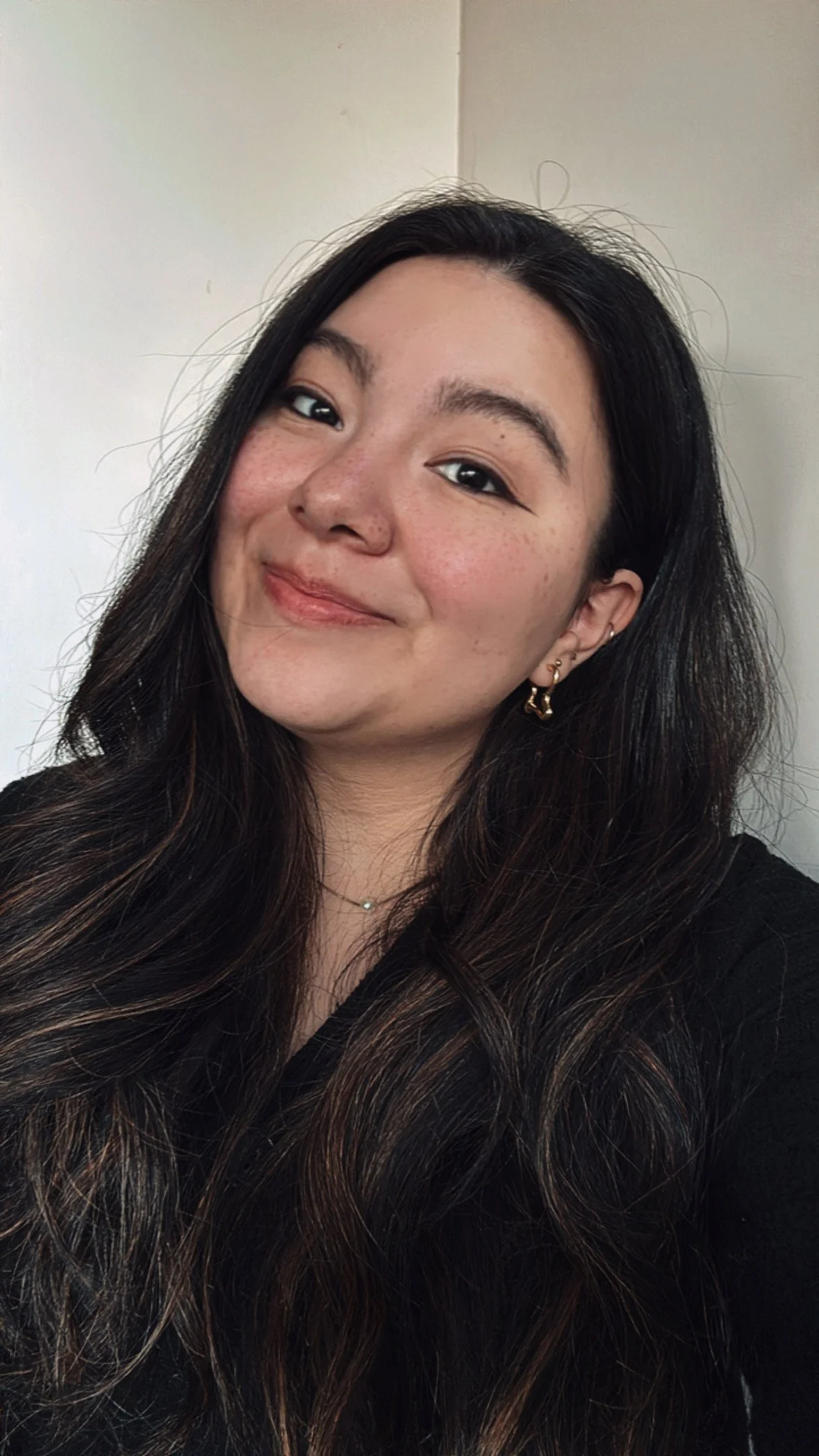 A young woman with long, wavy dark hair and gold earrings, smiling at the camera against a plain light-colored wall.