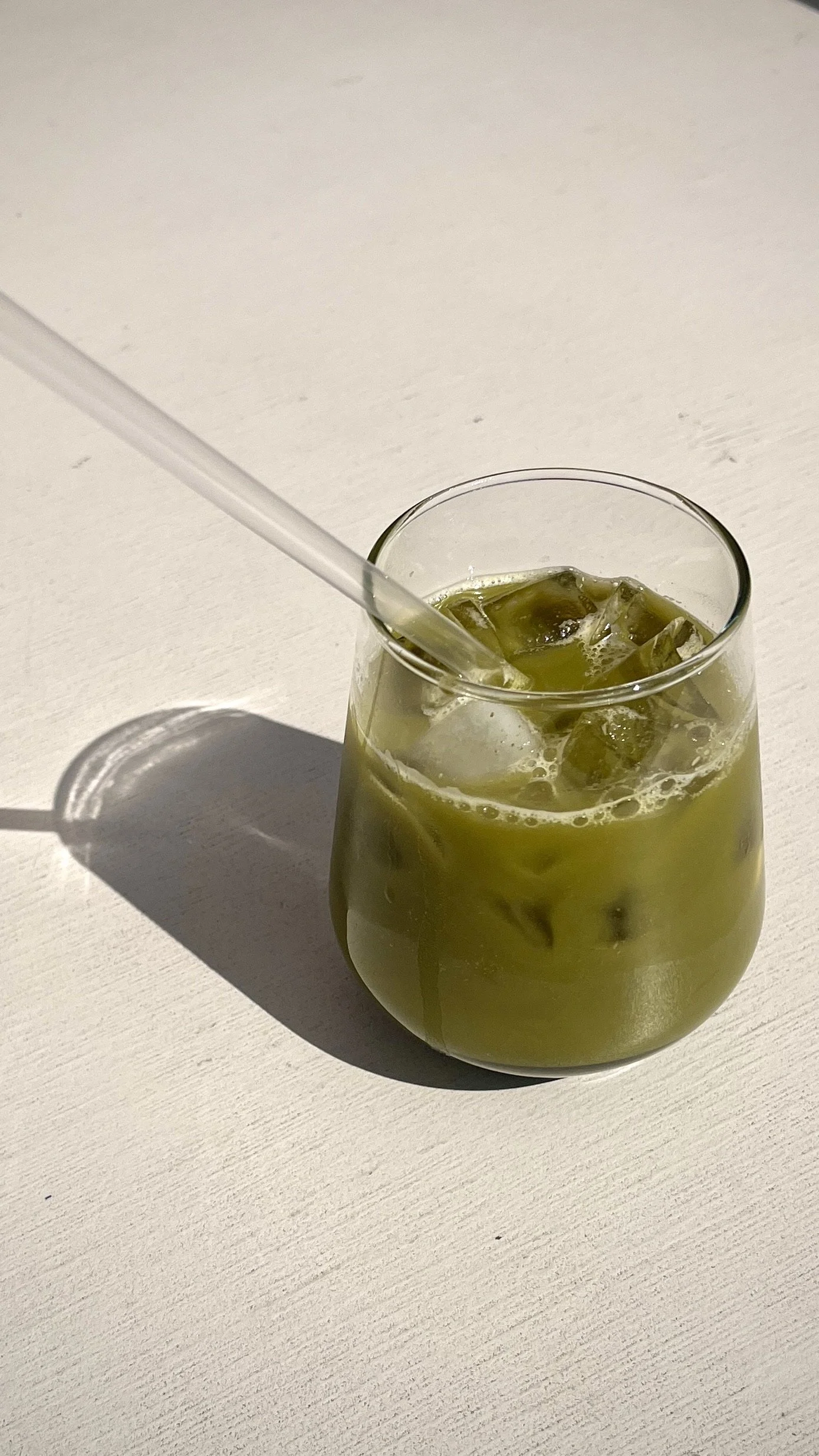 A glass of iced green tea with a white straw on a white surface.