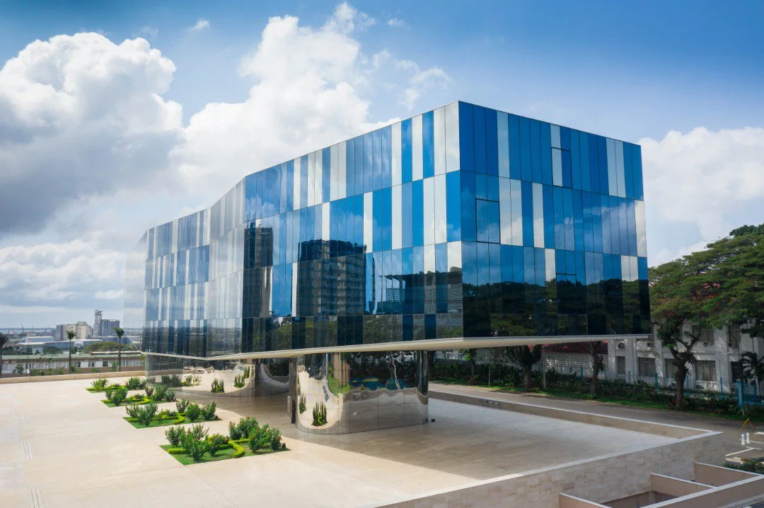 Bâtiment moderne avec une façade en verre bleue, reflet du ciel et des bâtiments environnants, dans une zone urbaine, sous un ciel partiellement nuageux.