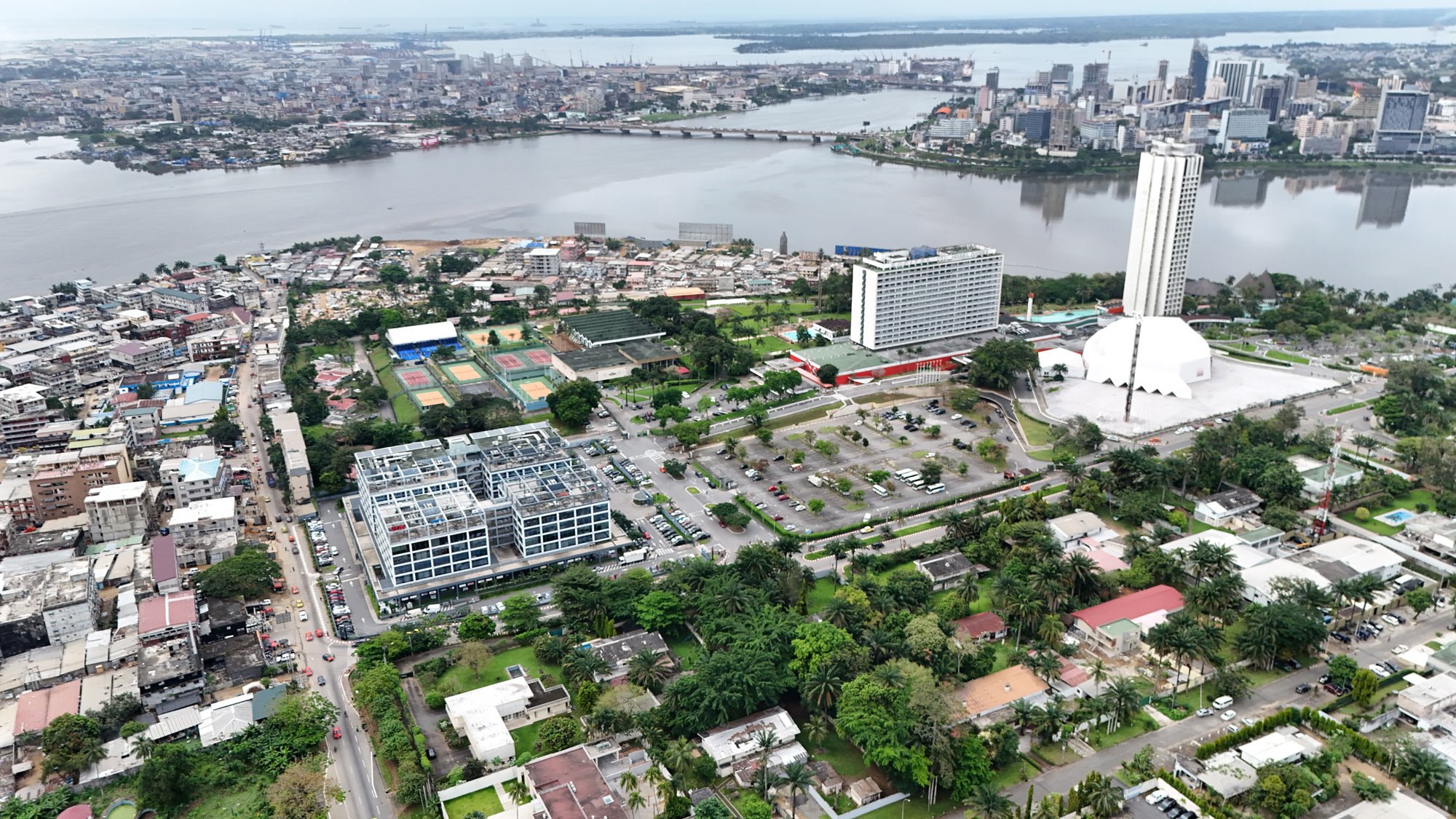 Vue aérienne d'une ville avec un fleuve, un parc avec terrains de tennis, des bâtiments modernes et résidentiels, et une grande structure blanche en forme de dôme.