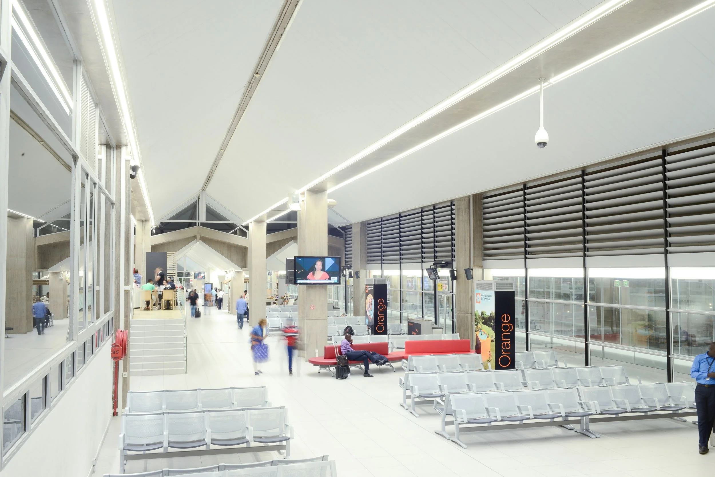 Salle d'attente d'un aéroport avec sièges blancs, écrans, et fenêtres panoramiques, quelques passagers assis ou marchant.