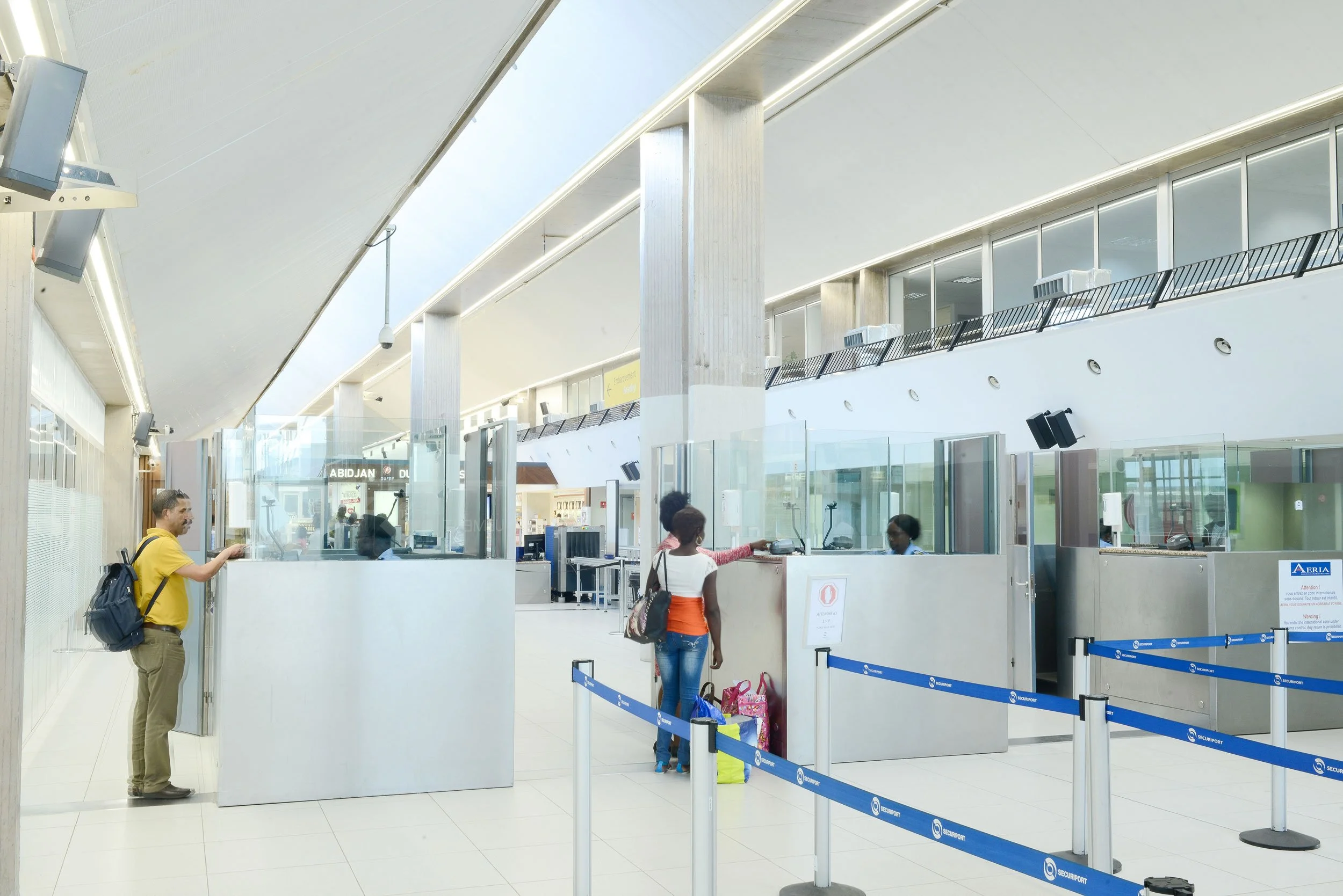 Intérieur d'un aéroport avec des comptoirs d'enregistrement et des passagers en attente, certains parlent avec le personnel, autres avec des sacs à dos ou bagages.