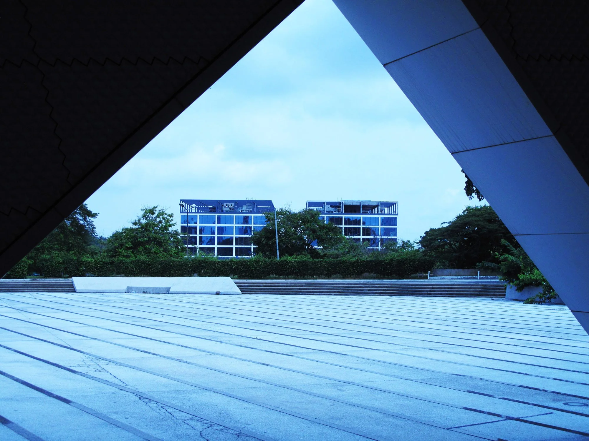 vue depuis une ouverture architecturale montrant deux bâtiments en verre derrière un espace pavé entouré de végétation.