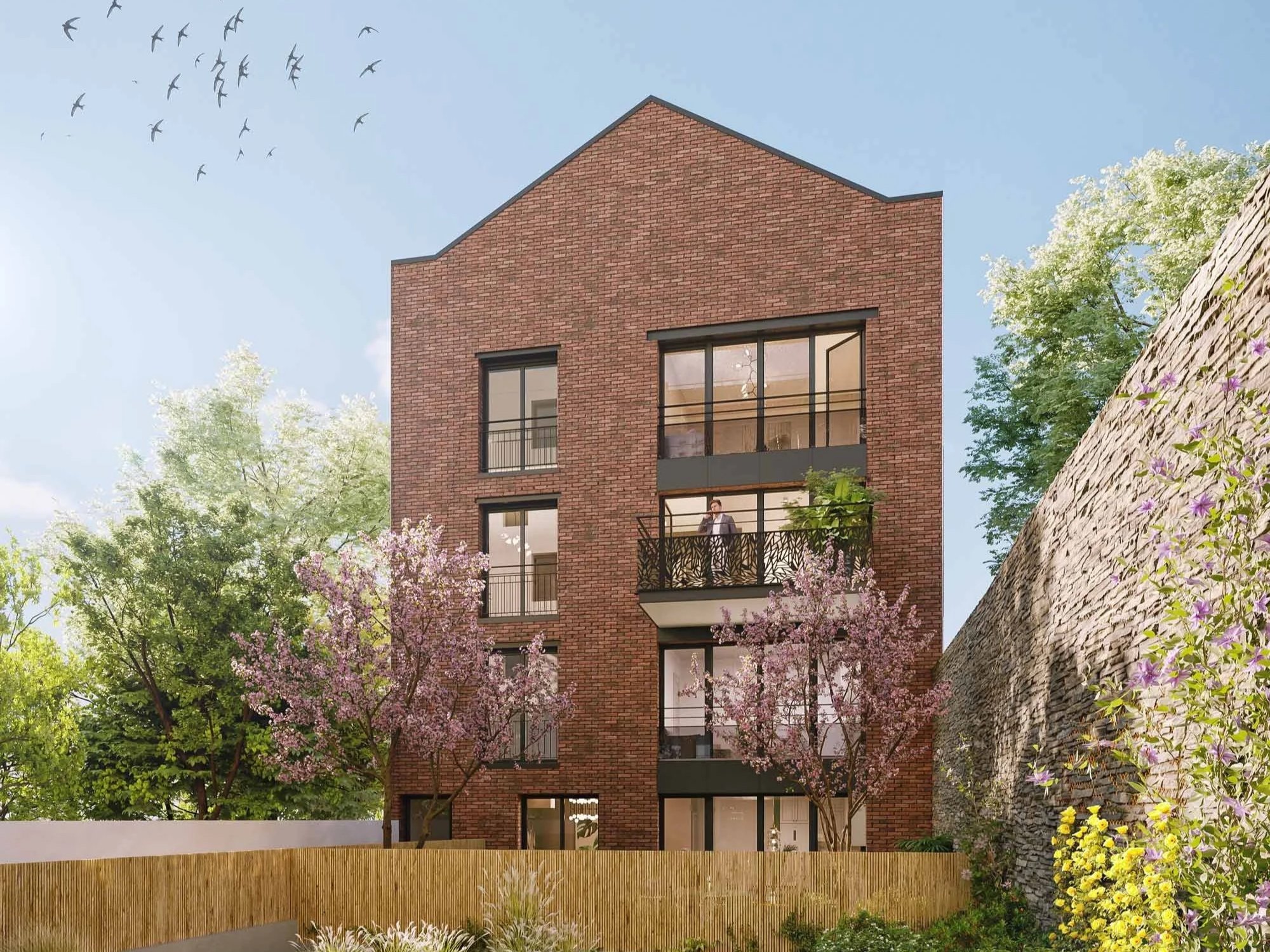 Immeuble résidentiel en briques rouges avec balcons, entouré d'arbres en fleurs et d'un mur de pierre, sous un ciel bleu avec des oiseaux