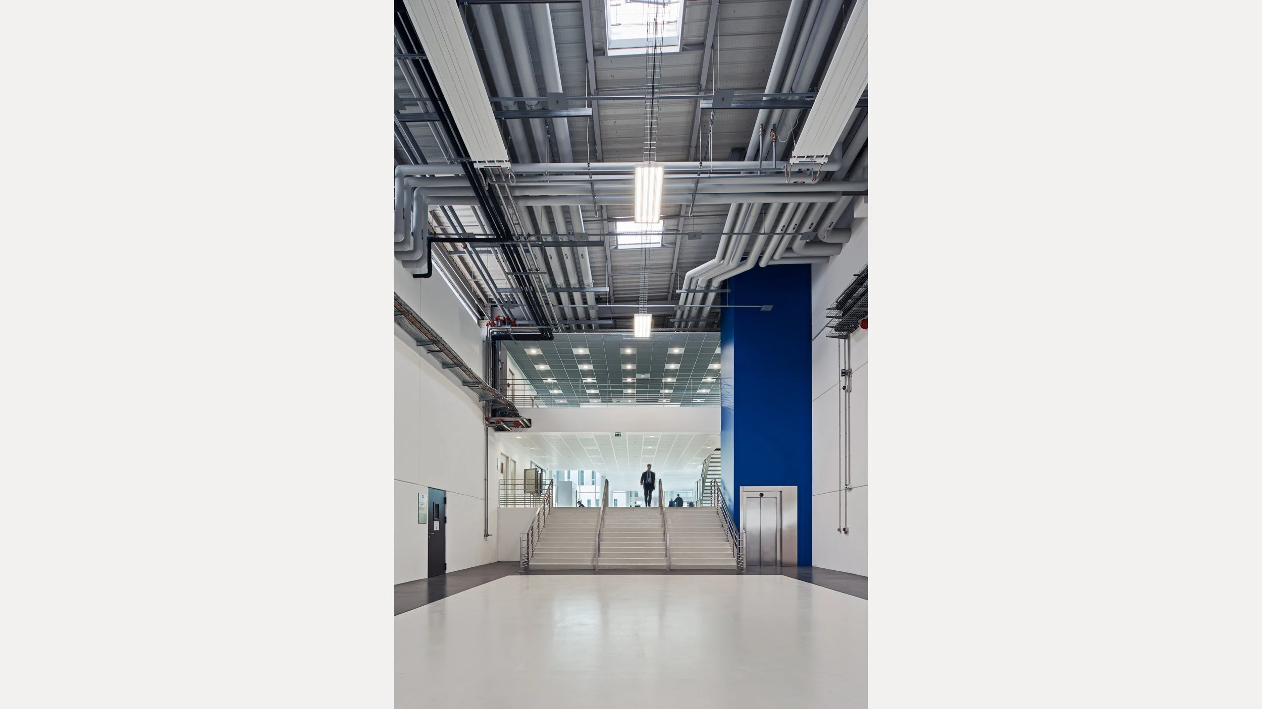 Intérieur d'un bâtiment moderne avec un escalier menant à un espace ouvert, un homme en costume au sommet des escaliers, et un plafond industriel avec pipes et luminaires.