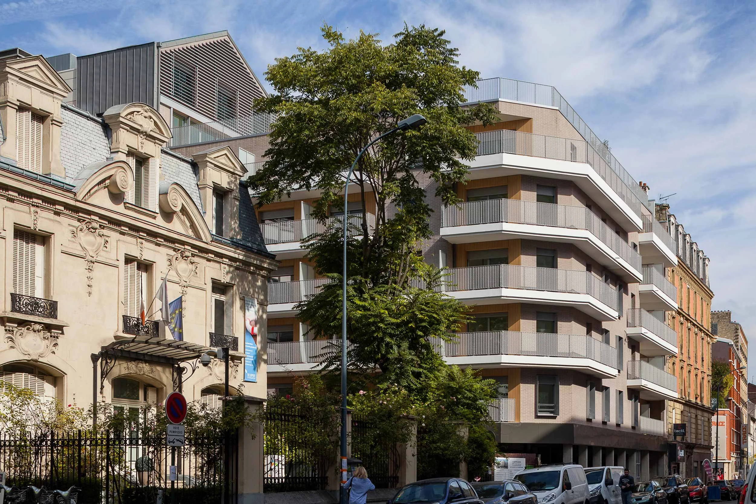Un bâtiment ancien en pierre avec des fenêtres à volets, un drapeau français et un drapeau européen, à côté d’un bâtiment moderne avec plusieurs balcons en cornet, sous un ciel bleu avec quelques nuages.