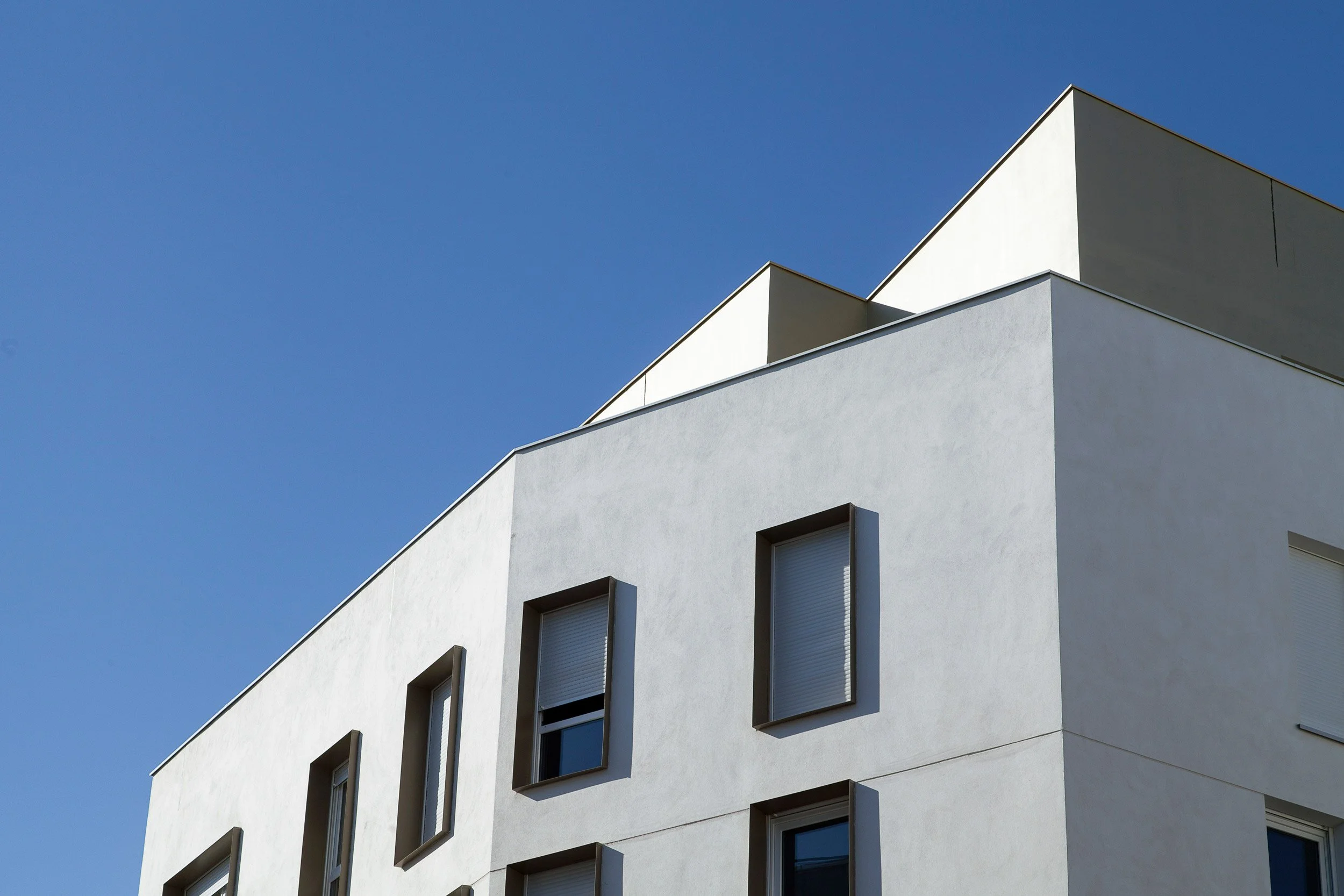 Vue d'un bâtiment moderne blanc avec plusieurs fenêtres et un toit en forme de pentagone sous un ciel clair.