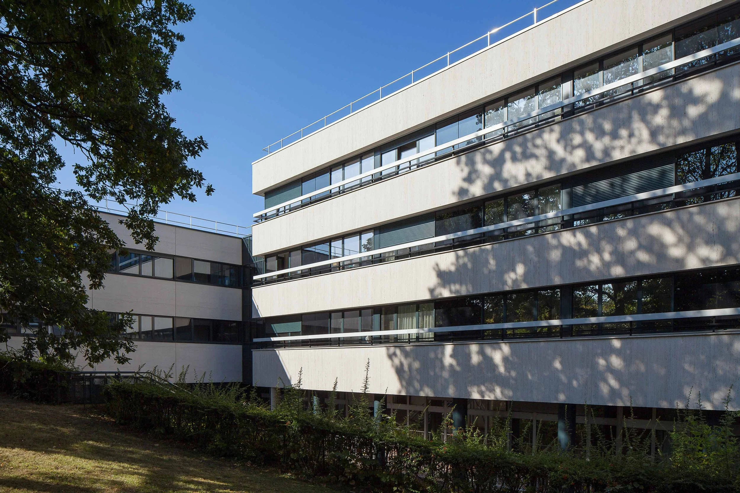 Bâtiment moderne avec plusieurs étages, grandes fenêtres et balustrades, entouré d'arbres et de végétation, sous un ciel bleu