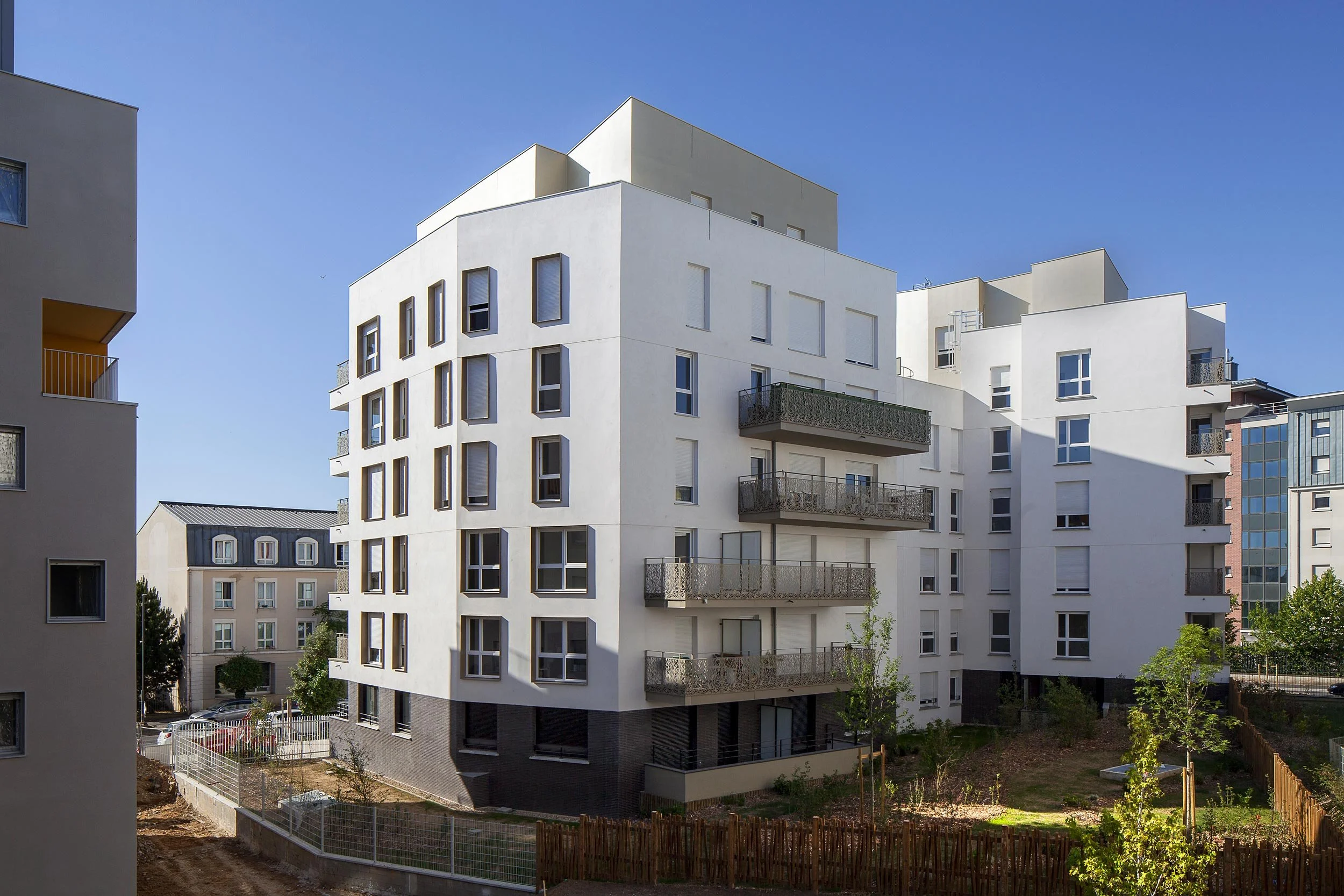 Immeuble résidentiel moderne blanc avec plusieurs balcons en fer forgé, situé dans un quartier urbain ensoleillé, avec un espace vert à l'avant et quelques jeunes arbres.