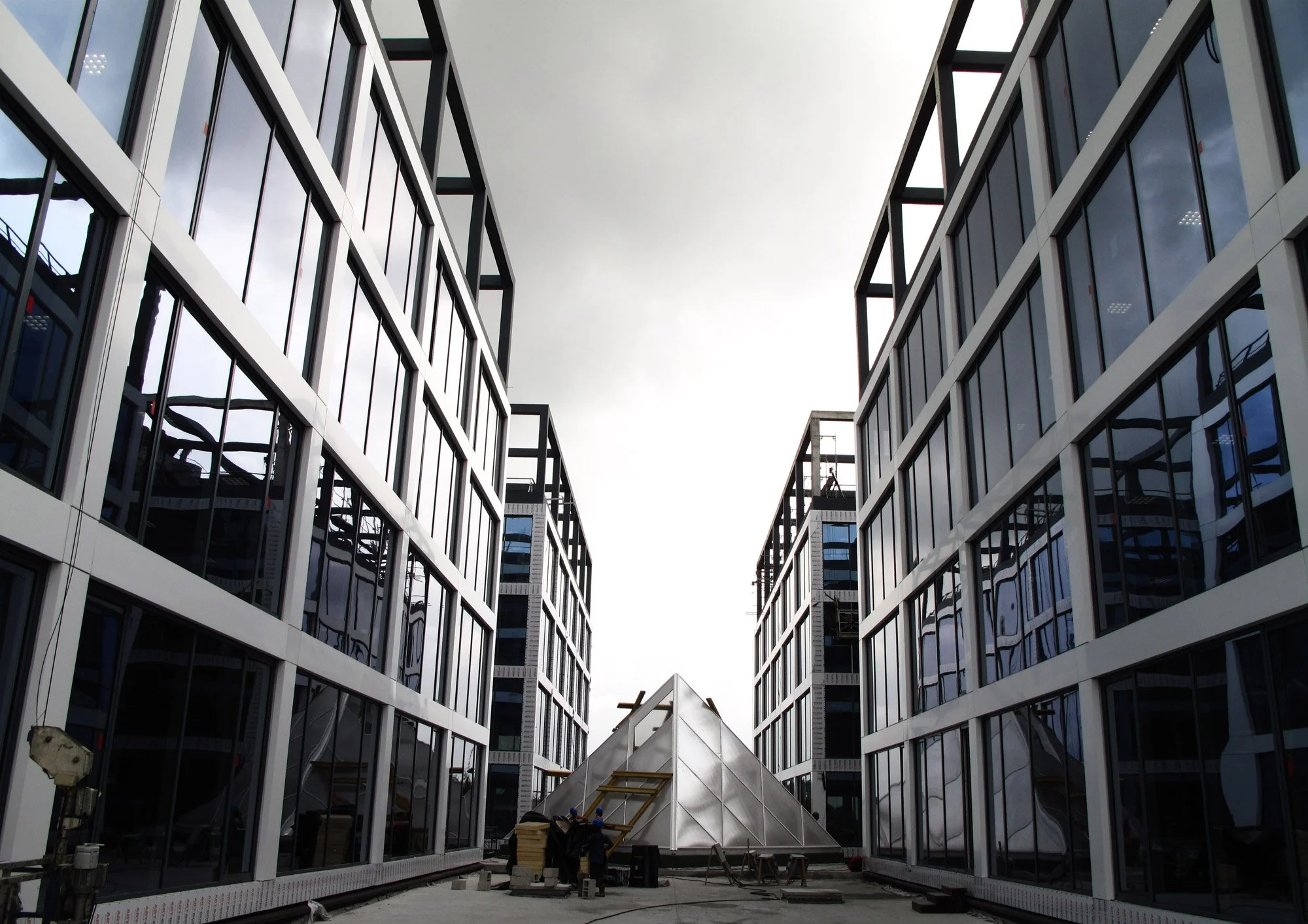 Vue d'un chantier avec des bâtiments modernes en verre et un toit pyramidal en métal au centre.