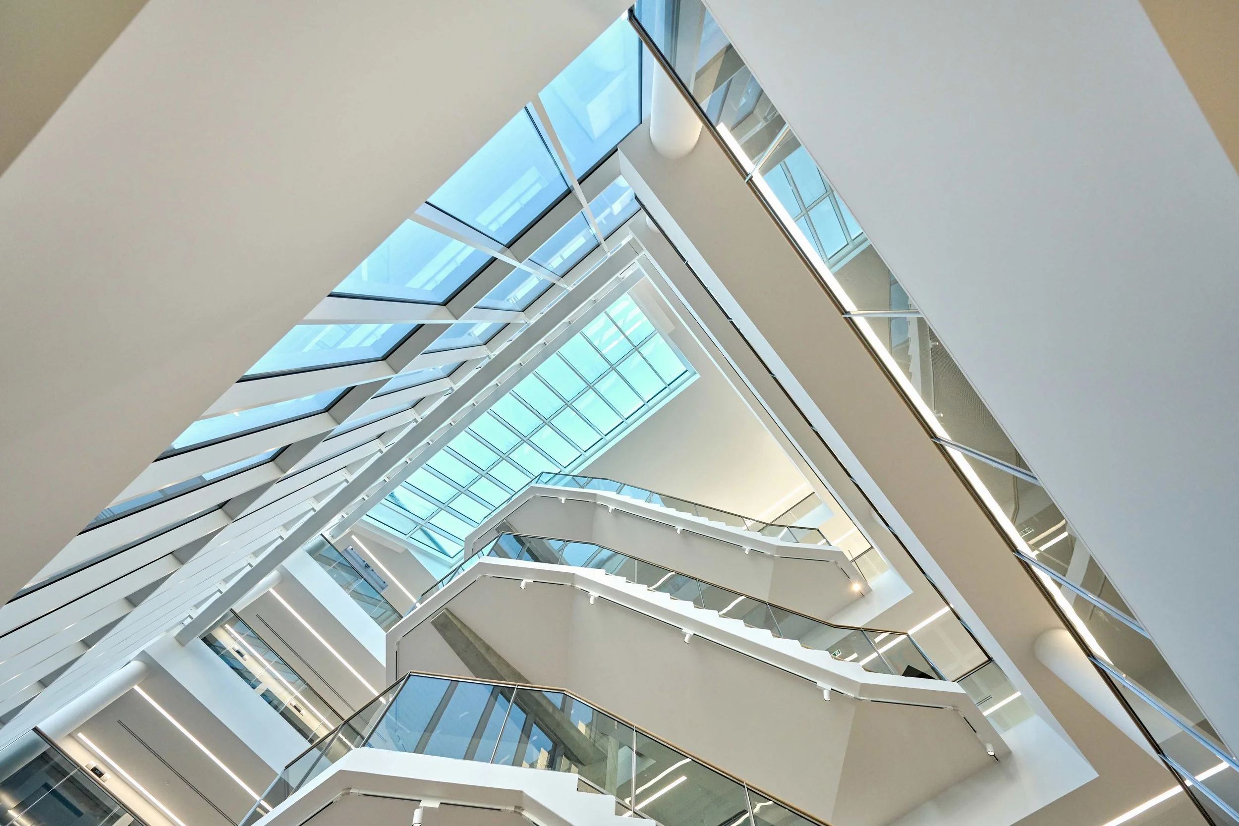Vue intérieure d'un bâtiment moderne avec plusieurs étages, escaliers en colimaçon, murs blancs, grandes fenêtres en verre et lumière naturelle.