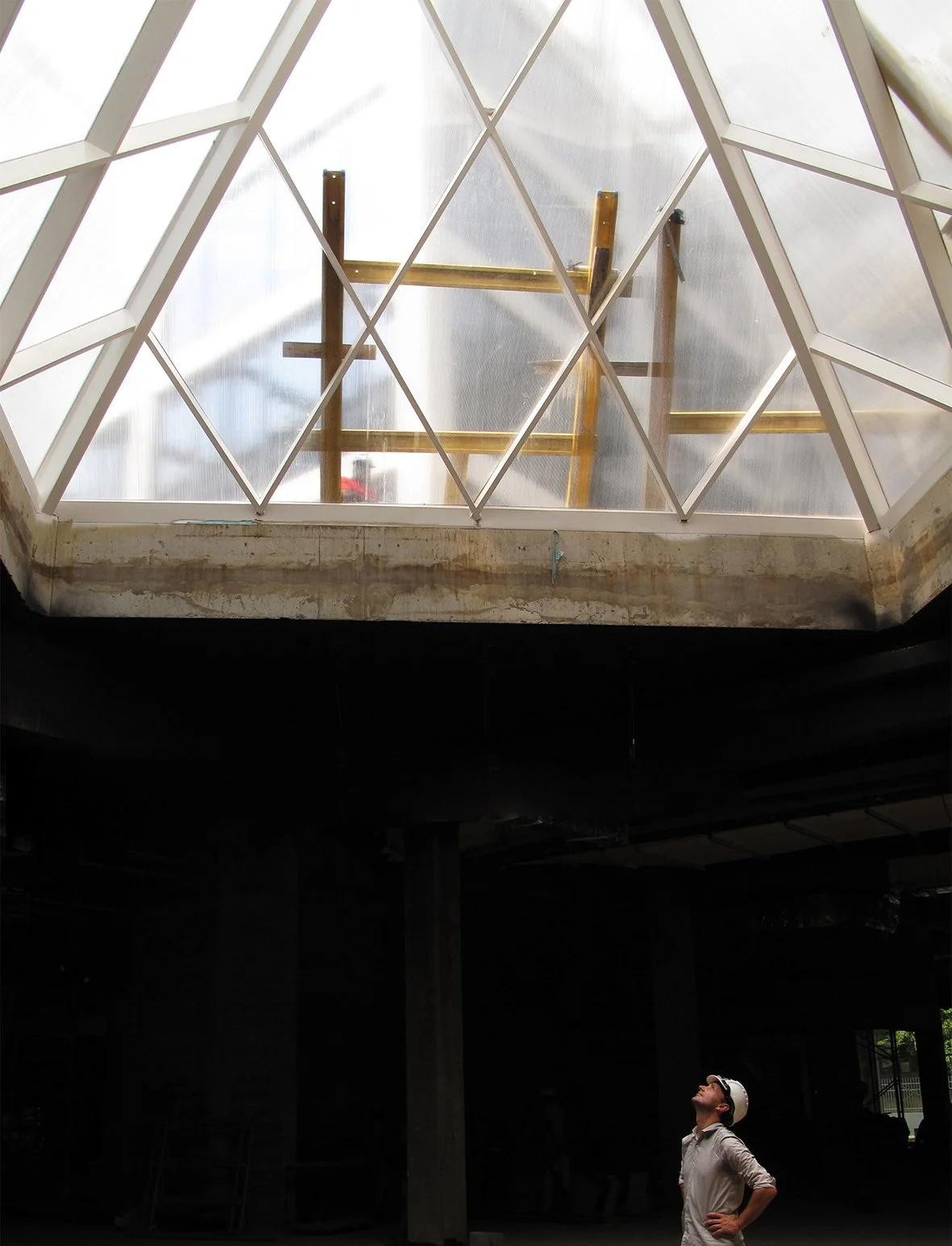 Un homme en casque de sécurité regarde vers le haut dans un bâtiment en construction, avec une grande lucarne en verre et une structure métallique visible au-dessus.