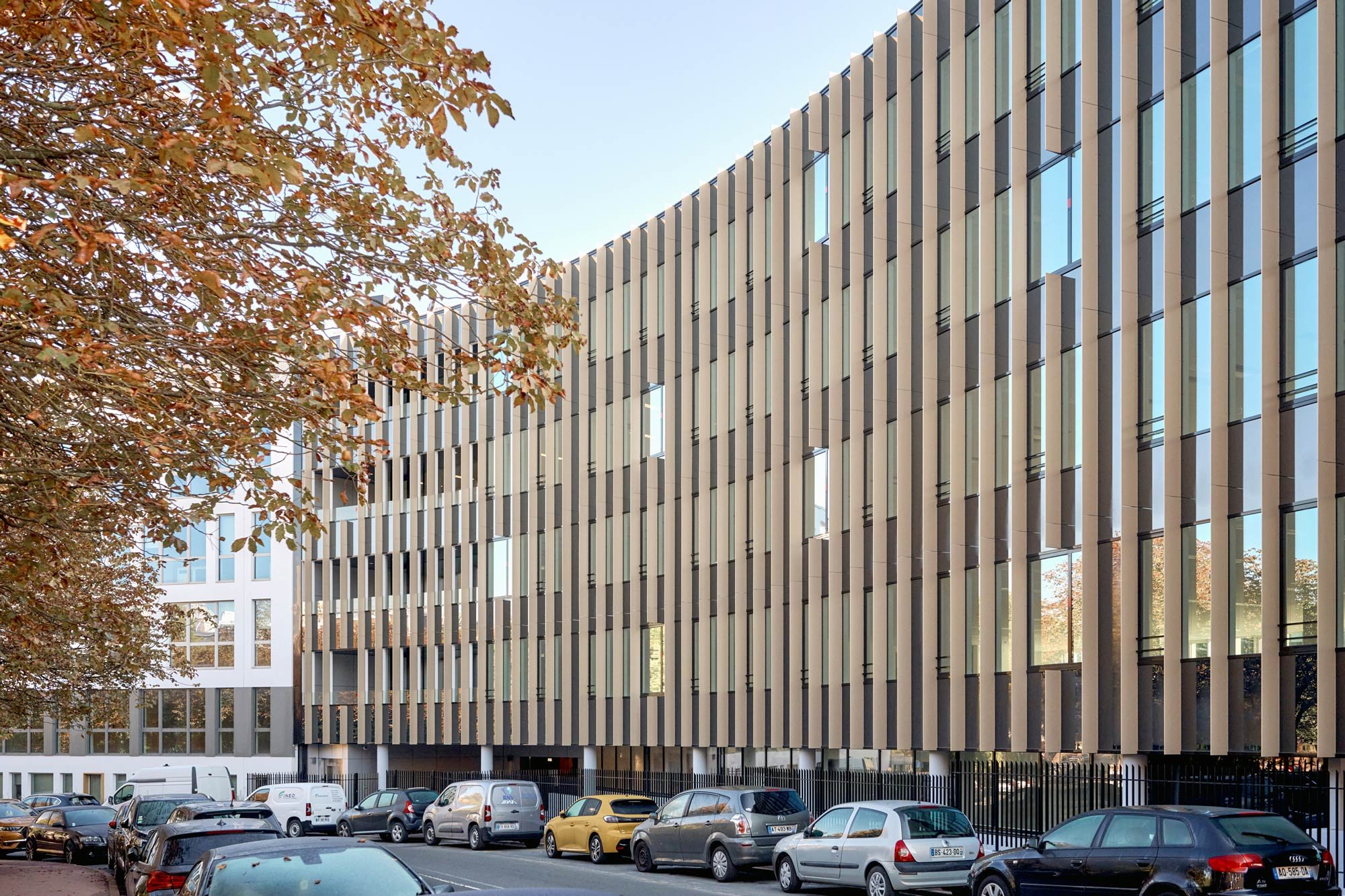 Façade moderne d'un bâtiment avec fenêtres comportant des volets verticaux en métal, vue depuis une rue avec plusieurs voitures garées. Un arbre avec des feuilles marron à gauche.