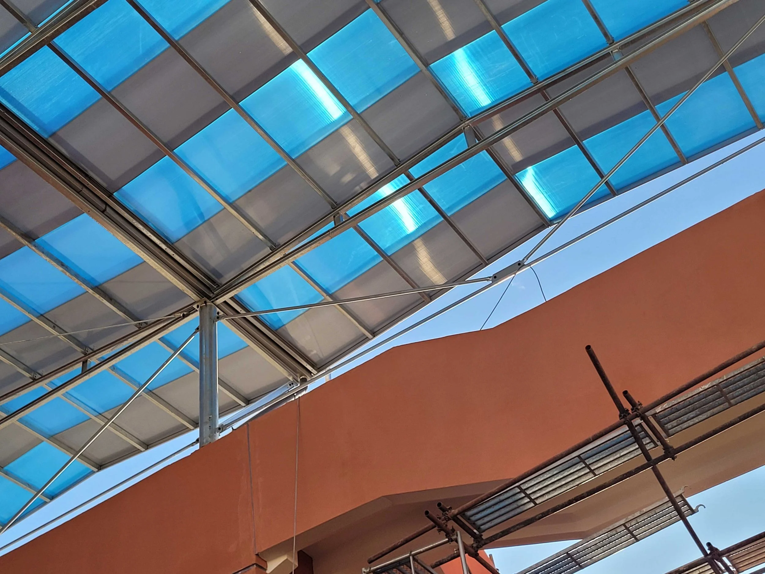 Vue en dessous d'un abri en verre avec structure métallique, ciel bleu en arrière-plan, et partie d'un bâtiment orange visible.