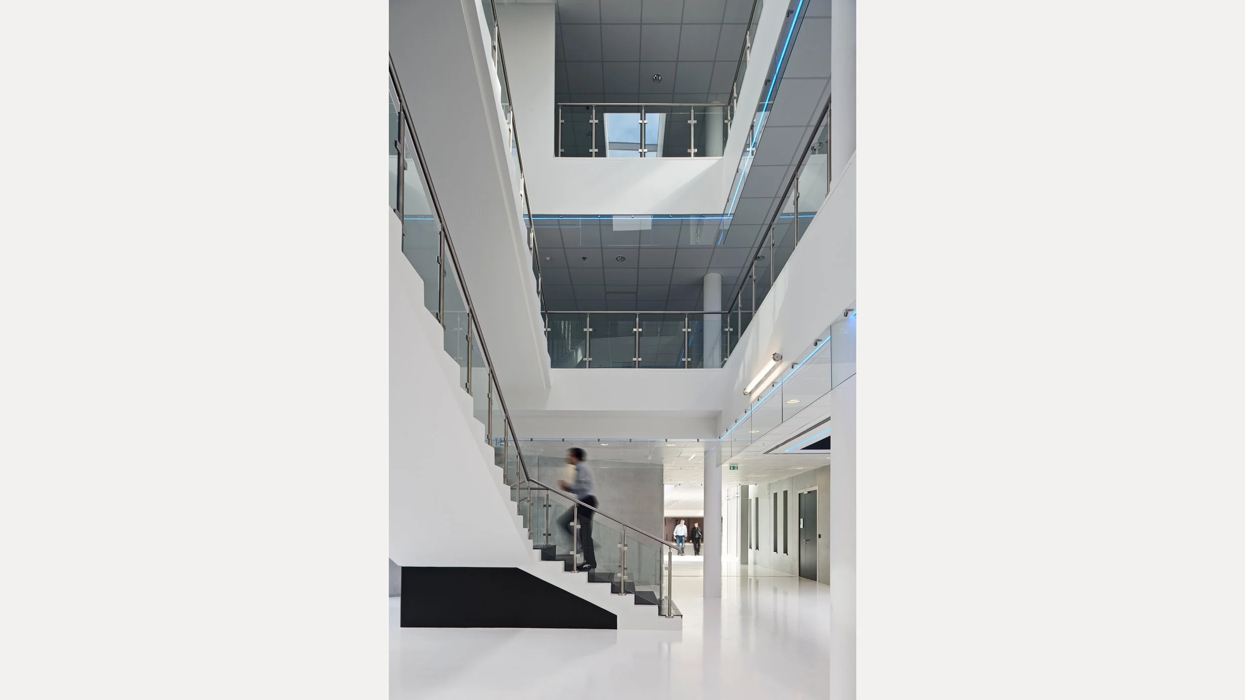 Interior moderne d'un bâtiment avec un escalier en colimaçon, plusieurs étages visibles, des balustrades en verre, et des personnes marchant dans le couloir lumineux.