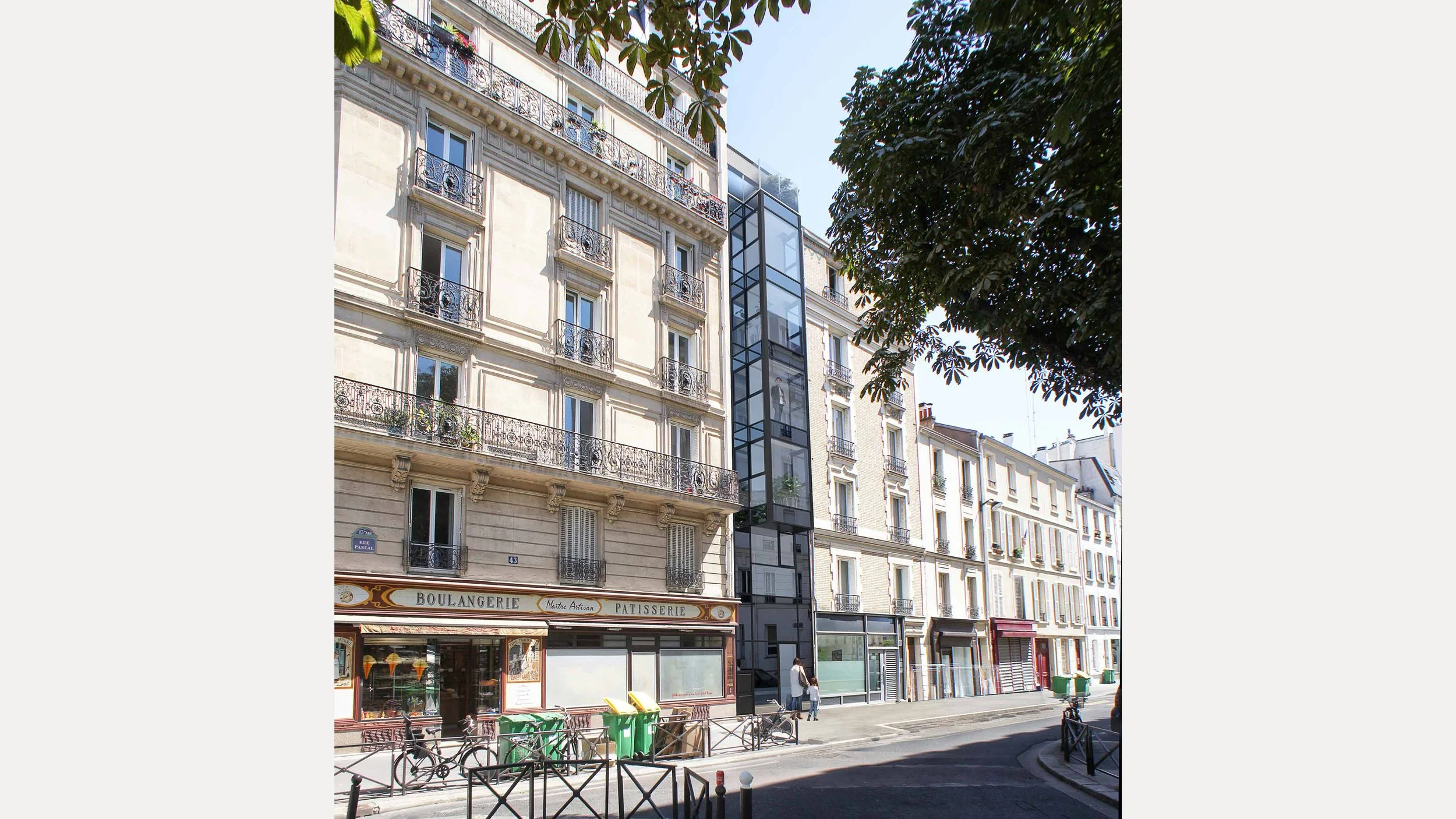 Rue avec immeubles haussmanniens, un ascenseur extérieur en verre, une boulangerie, des piétons, des vélos, et des poubelles vertes