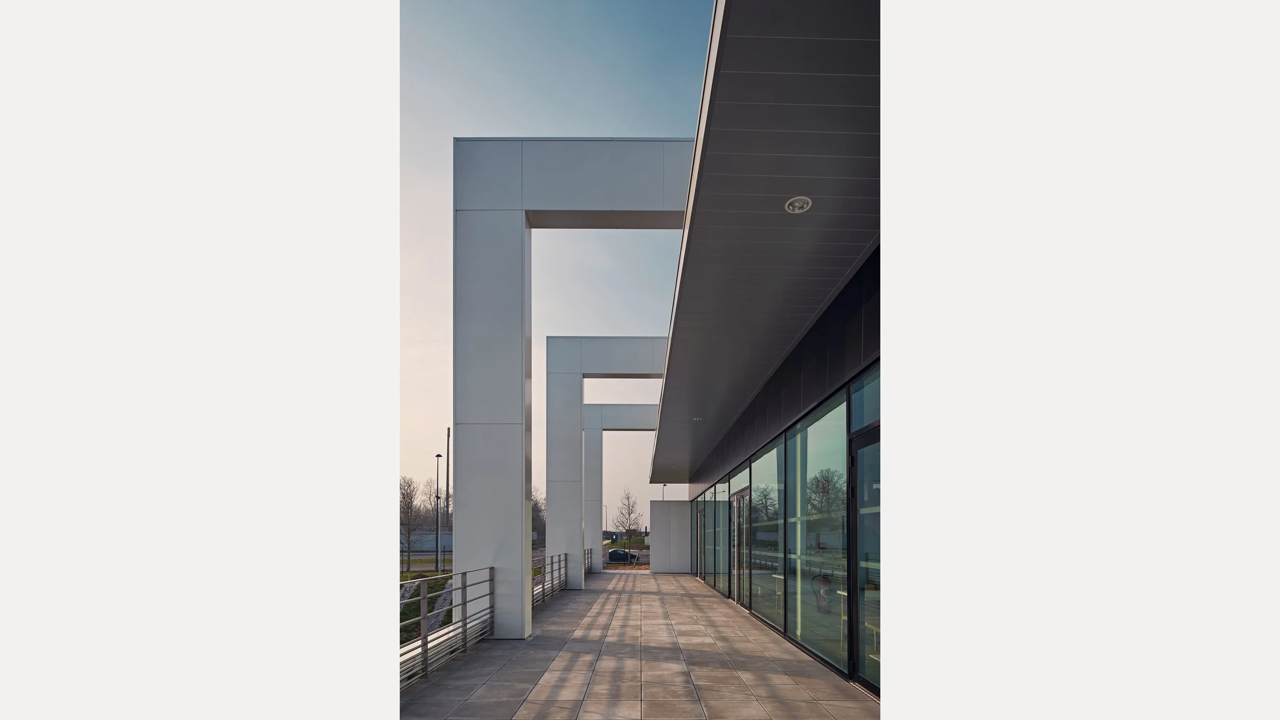 Bâtiment moderne avec de grandes fenêtres en verre et une terrasse en pierre, sous un ciel clair, avec des structures architecturales géométriques en blanc et noir.