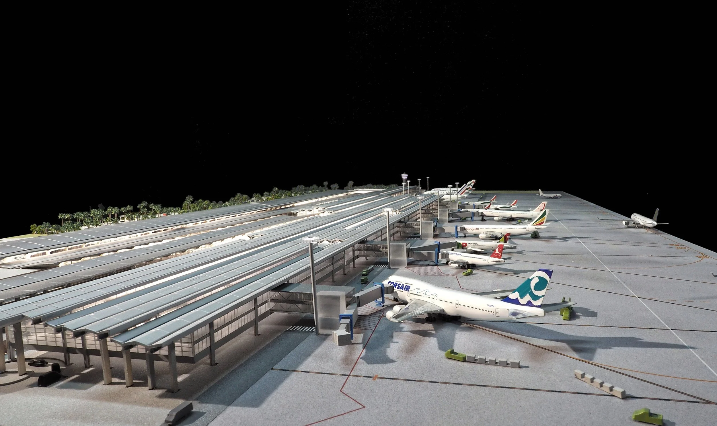Modèle réduit d'un aéroport avec plusieurs avions stationnés à la porte d'embarquement, un terminal en arrière-plan, et un ciel noir.