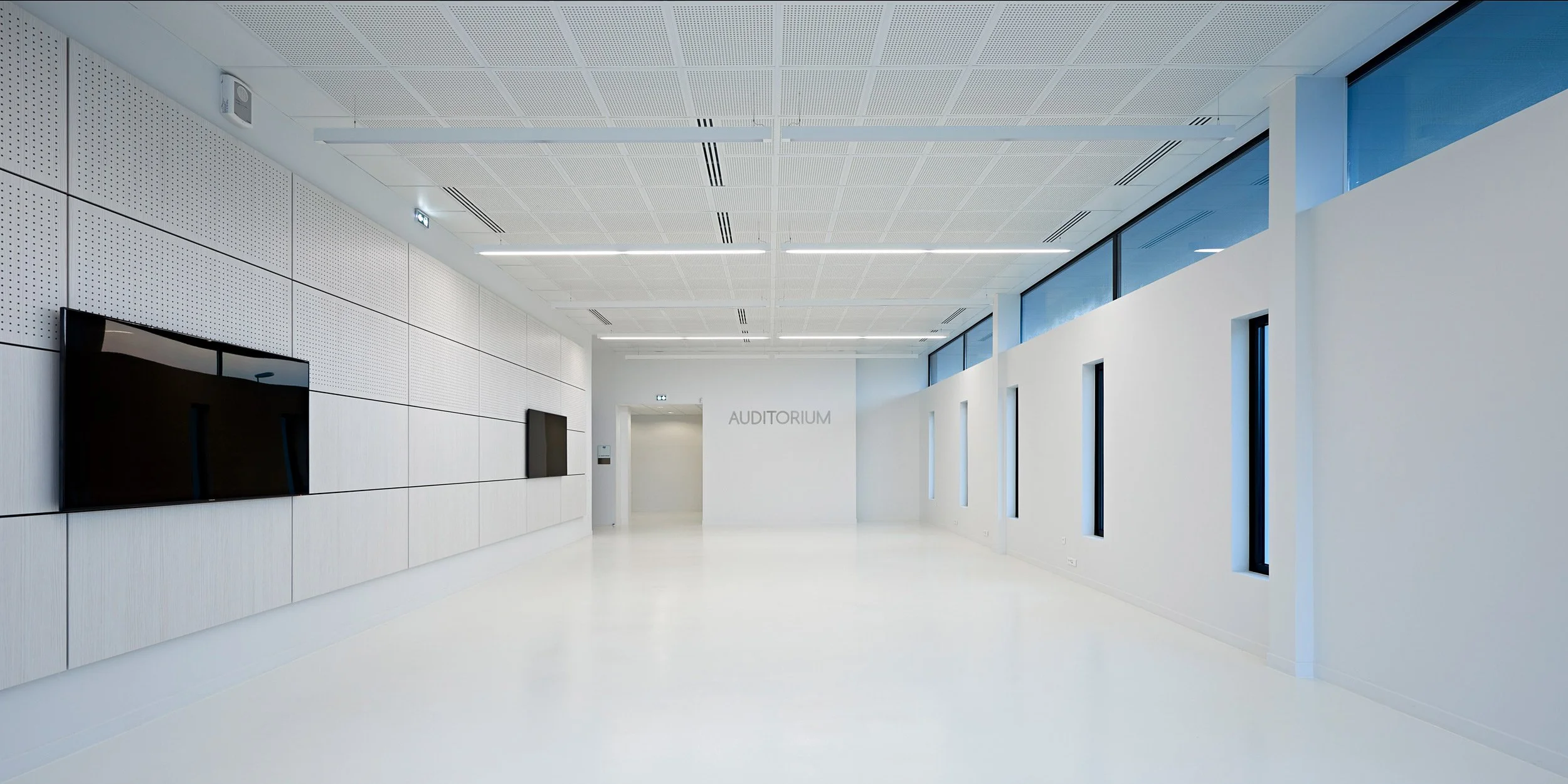 Une grande salle blanche lumineuse avec des murs et un sol blanc, plusieurs fenêtres alignées, un panneau indiquant 'AUDITORIUM' et deux écrans plats noirs fixés au mur.
