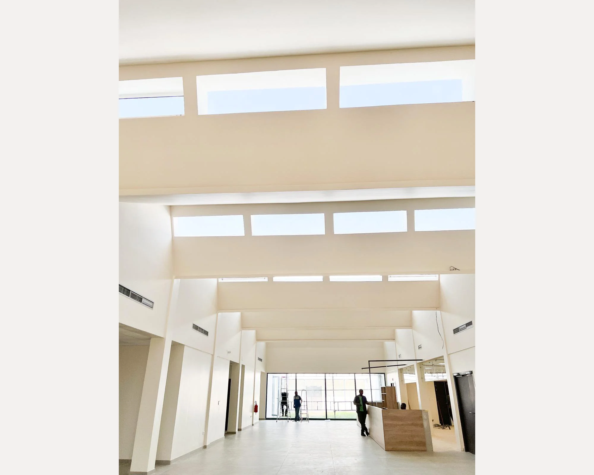 Hall moderne avec haut plafond, fenêtres en haut, réception en bois clair et personnes à l'accueil.