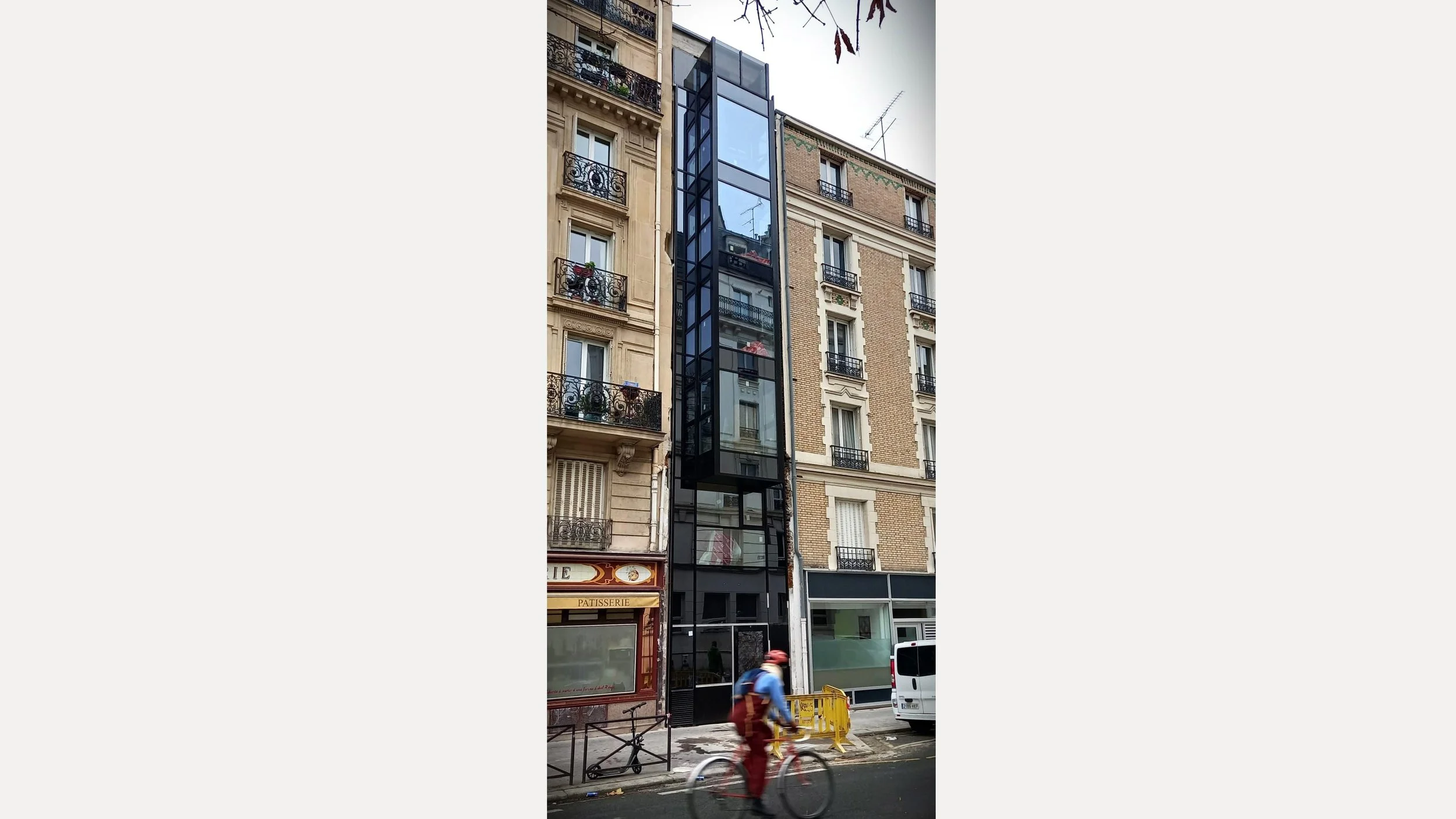 Un bâtiment moderne avec une façade en verre noir se trouvant entre deux bâtiments classiques parisiens. Au rez-de-chaussée, une pâtisserie et des fenêtres.