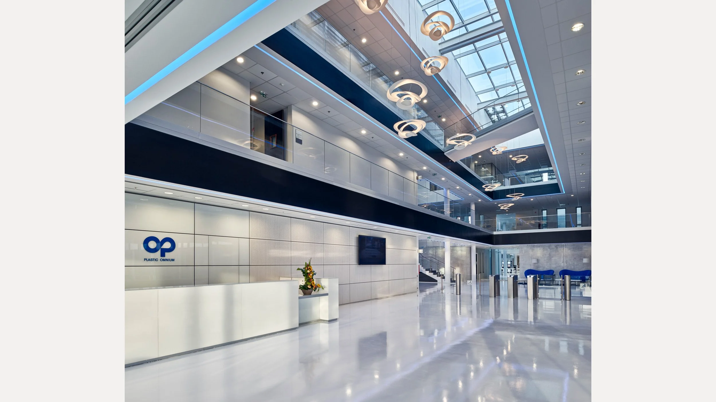 Véranda moderne d'une entreprise avec un comptoir blanc, un logo 'Plastic Omnium' en arrière-plan, un écran plat, des plantes décoratives, un sol en marbre brillant, et des plafonds avec des luminaires circulaires suspendus, tout dans des couleurs blanc, bleu et gris.