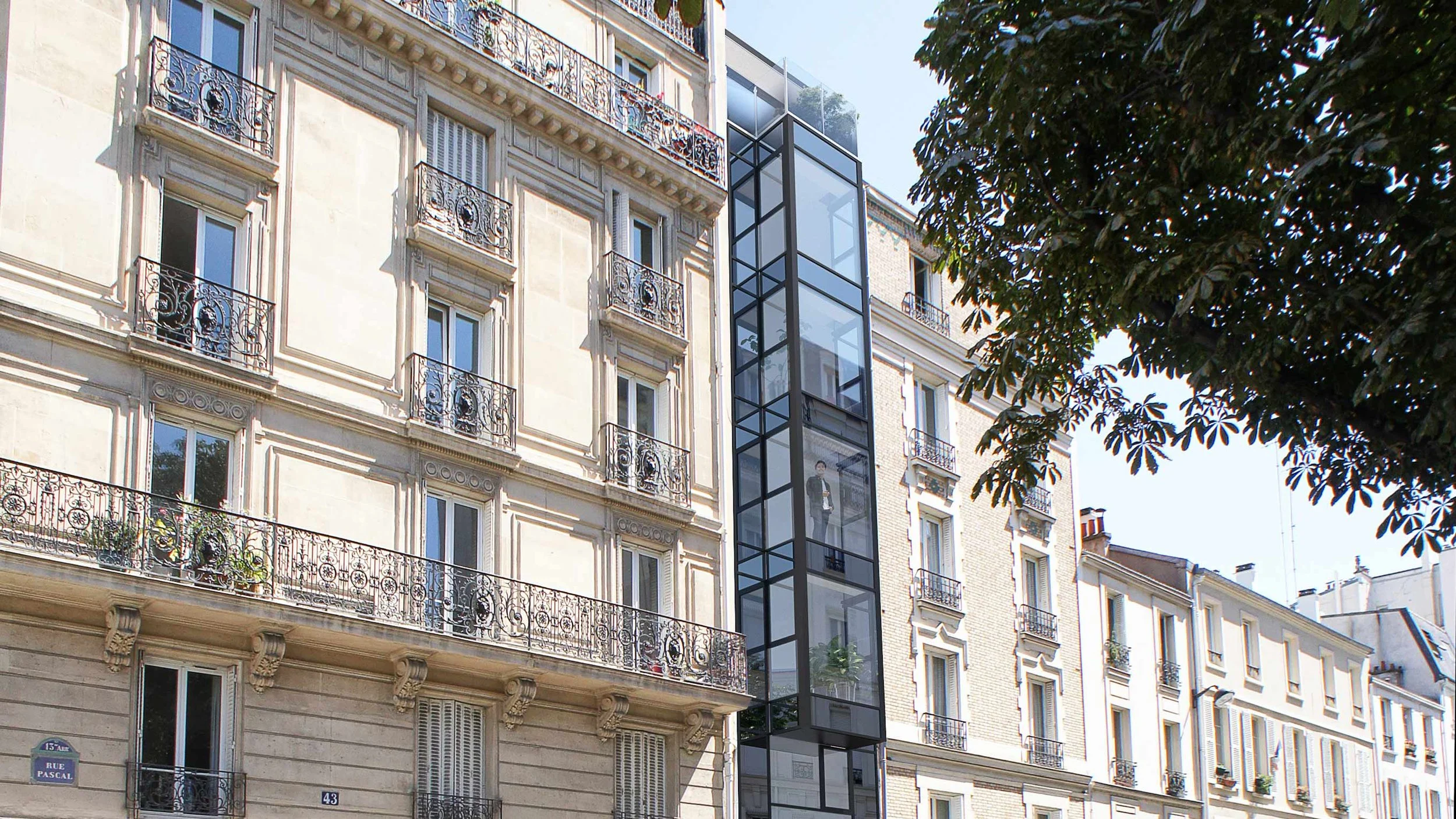 Une façade d'immeuble haussmannien avec des balcons en fer forgé et une échelle d'accès pour un ascenseur extérieur en verre et métal noir, avec un arbre à droite.