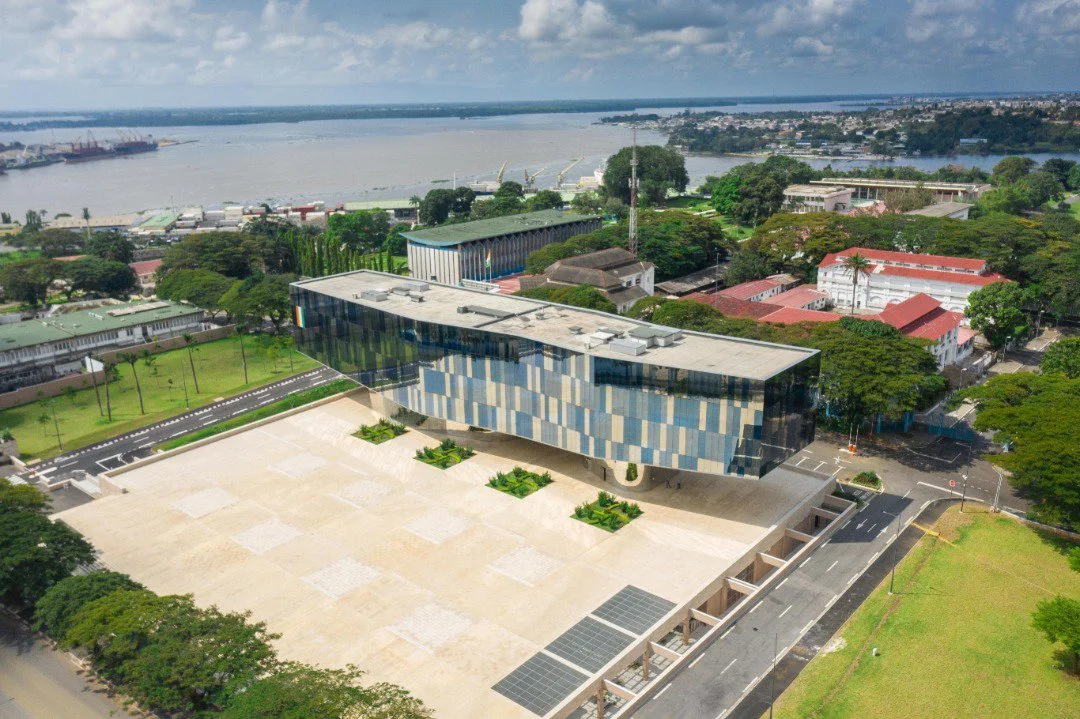 Vue aérienne d'un bâtiment moderne avec une façade en mosaïque bleue, situé dans un parc en zone urbaine près d'une rivière.