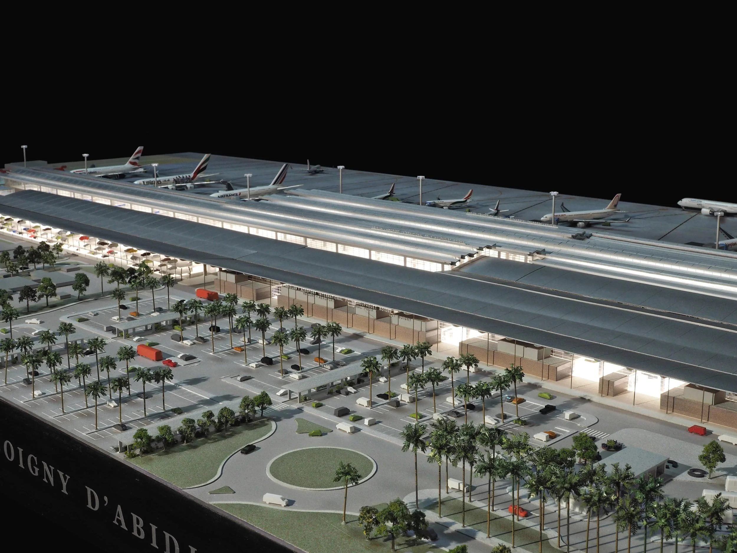 Modèle réduit d'un aéroport avec plusieurs avions stationnés, un parking avec voitures et arbres, et un bâtiment principal.
