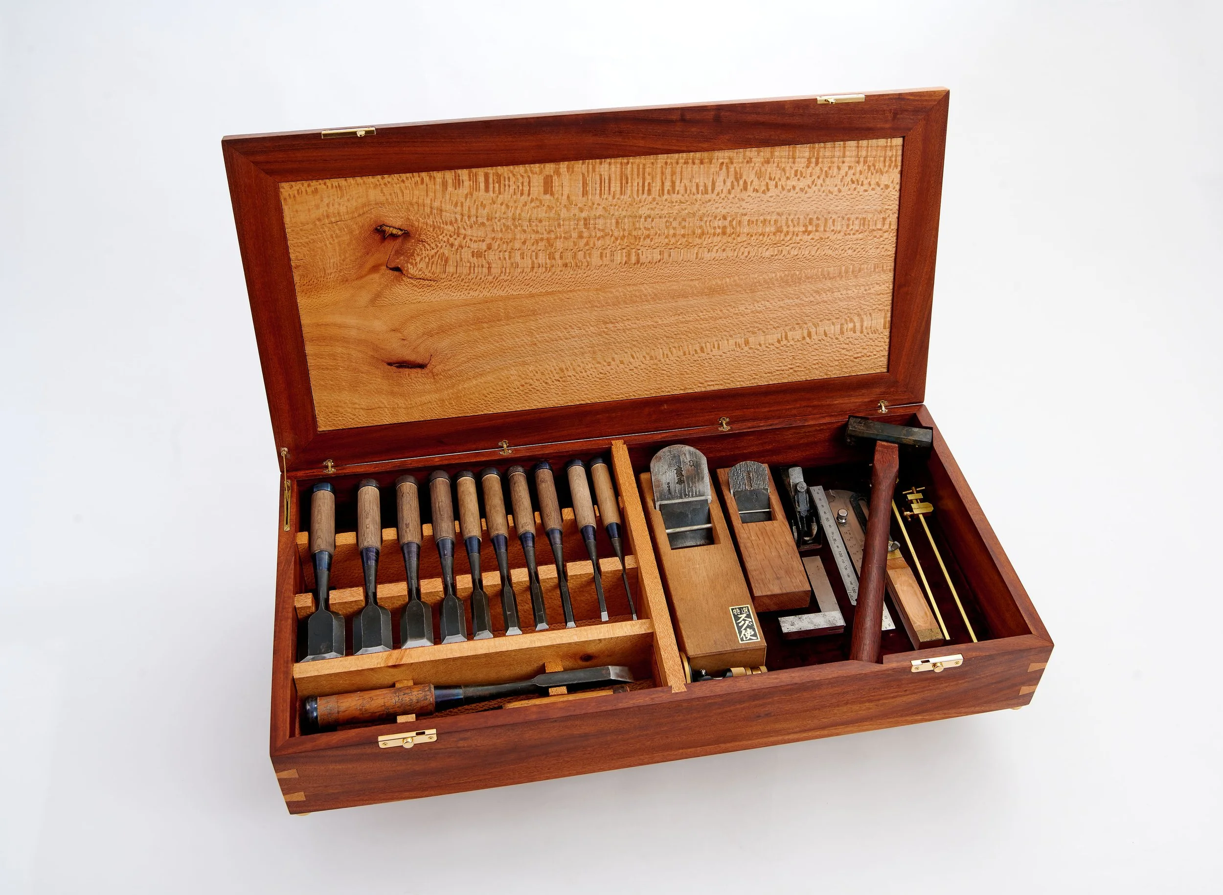 A wooden box with a hinged lid open revealing various woodworking tools inside, including chisels, files, and metal tools, set against a plain white background.