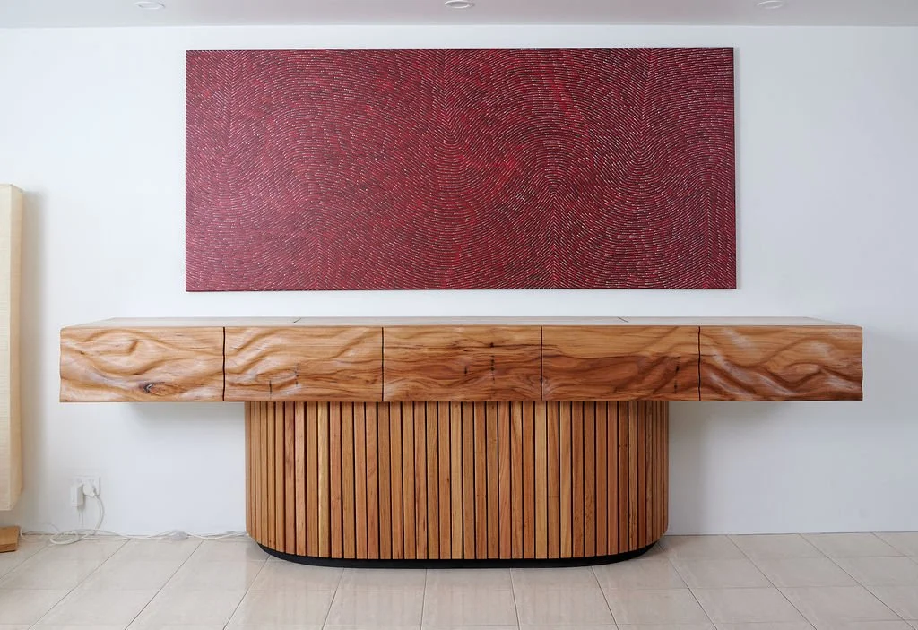 Wooden sideboard with vertical slats and a light wood top, with a large red textured artwork hanging above it, against a white wall.