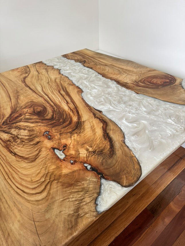 A wooden table with a natural, irregular edge, featuring swirling wood grain patterns and a pearl white resin river.