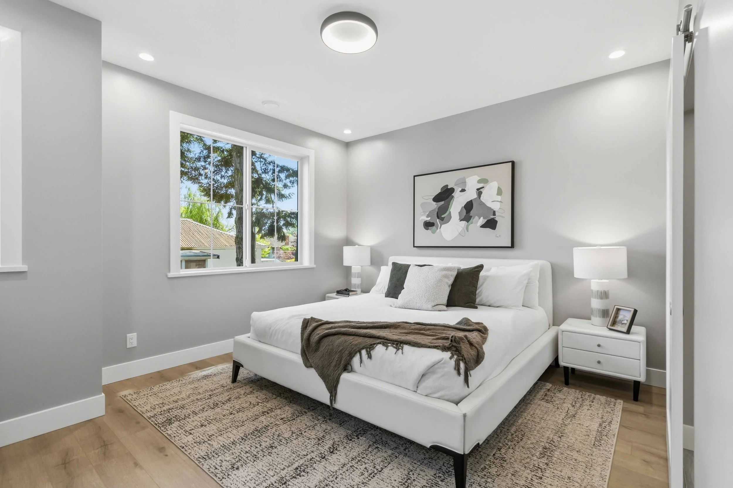 A modern bedroom with a white bed, gray pillows, a brown blanket, nightstands with lamps, large window showing trees outside, abstract artwork on the wall, and hardwood flooring.